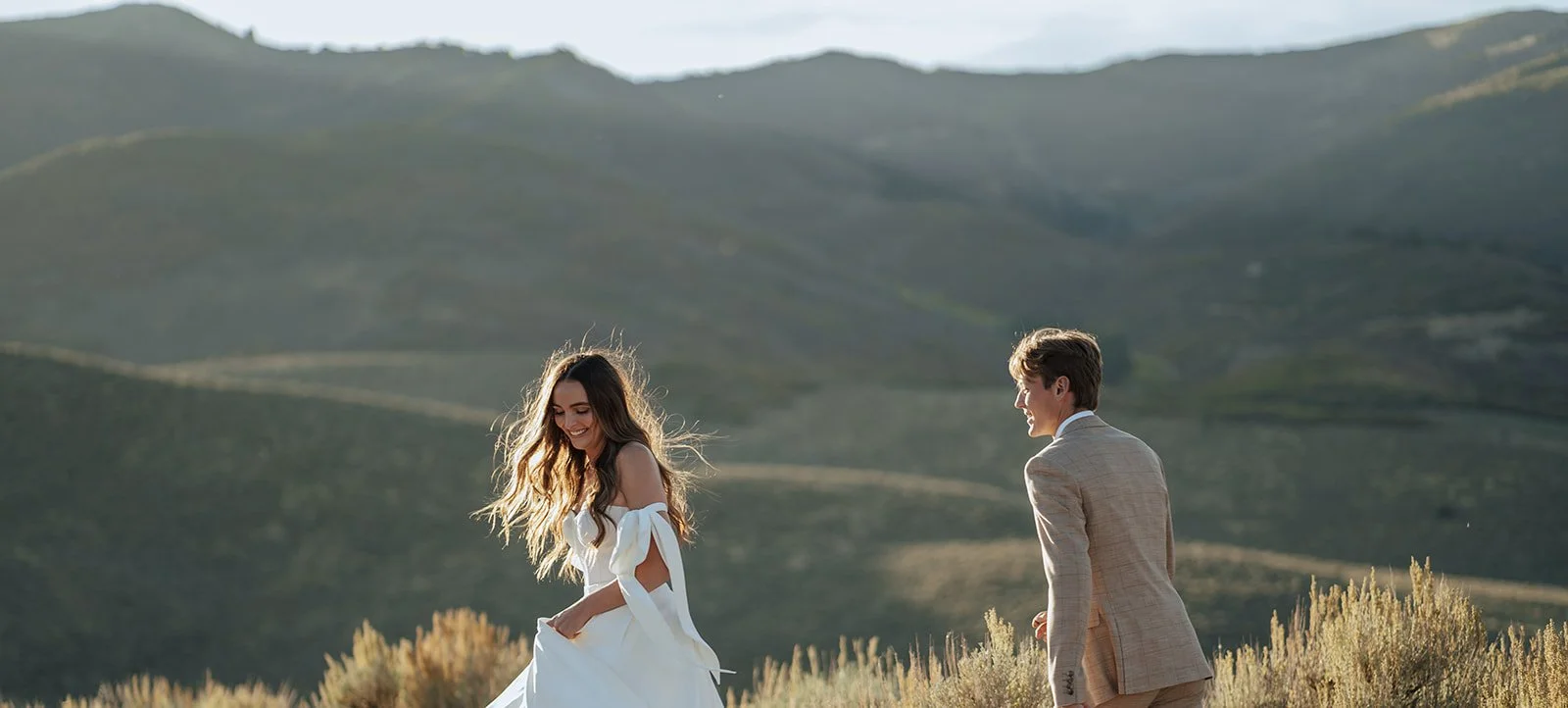 A bride and groom in a wedding gown and a tan suit stand in a meadow with mountainous peaks behind them - photography by Poise and Ivy Imagery