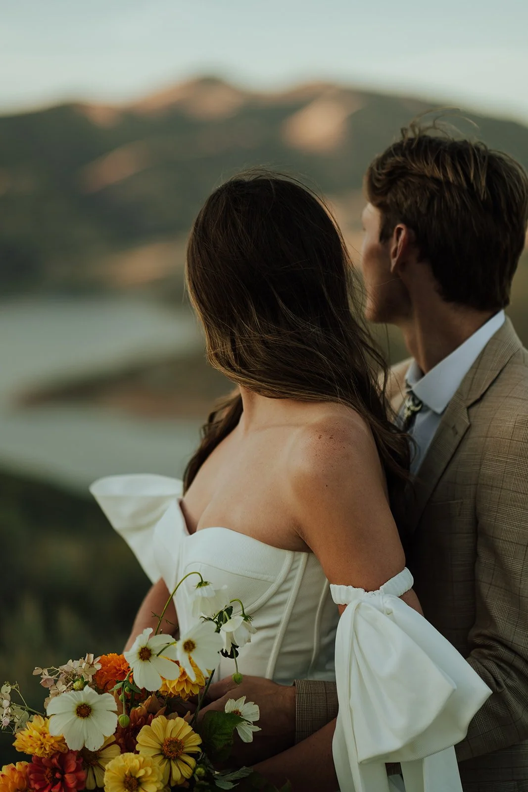 A bride and groom in a wedding gown and a tan suit stand in a meadow with mountains and a lake behind them - photography by Poise and Ivy Imagery
