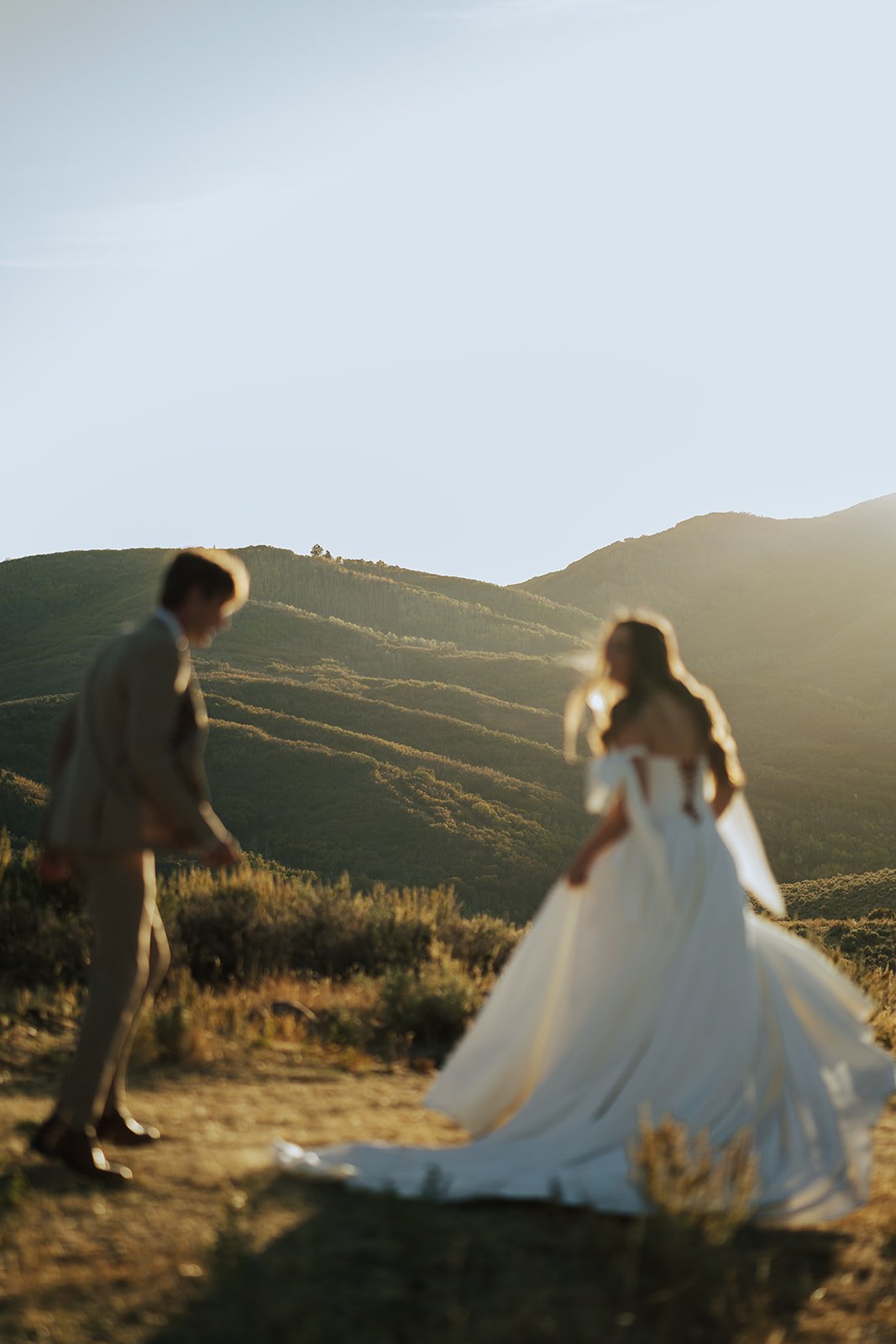 A bride and groom in a wedding gown and a tan suit stand in a meadow with mountainous peaks behind them - photography by Poise and Ivy Imagery