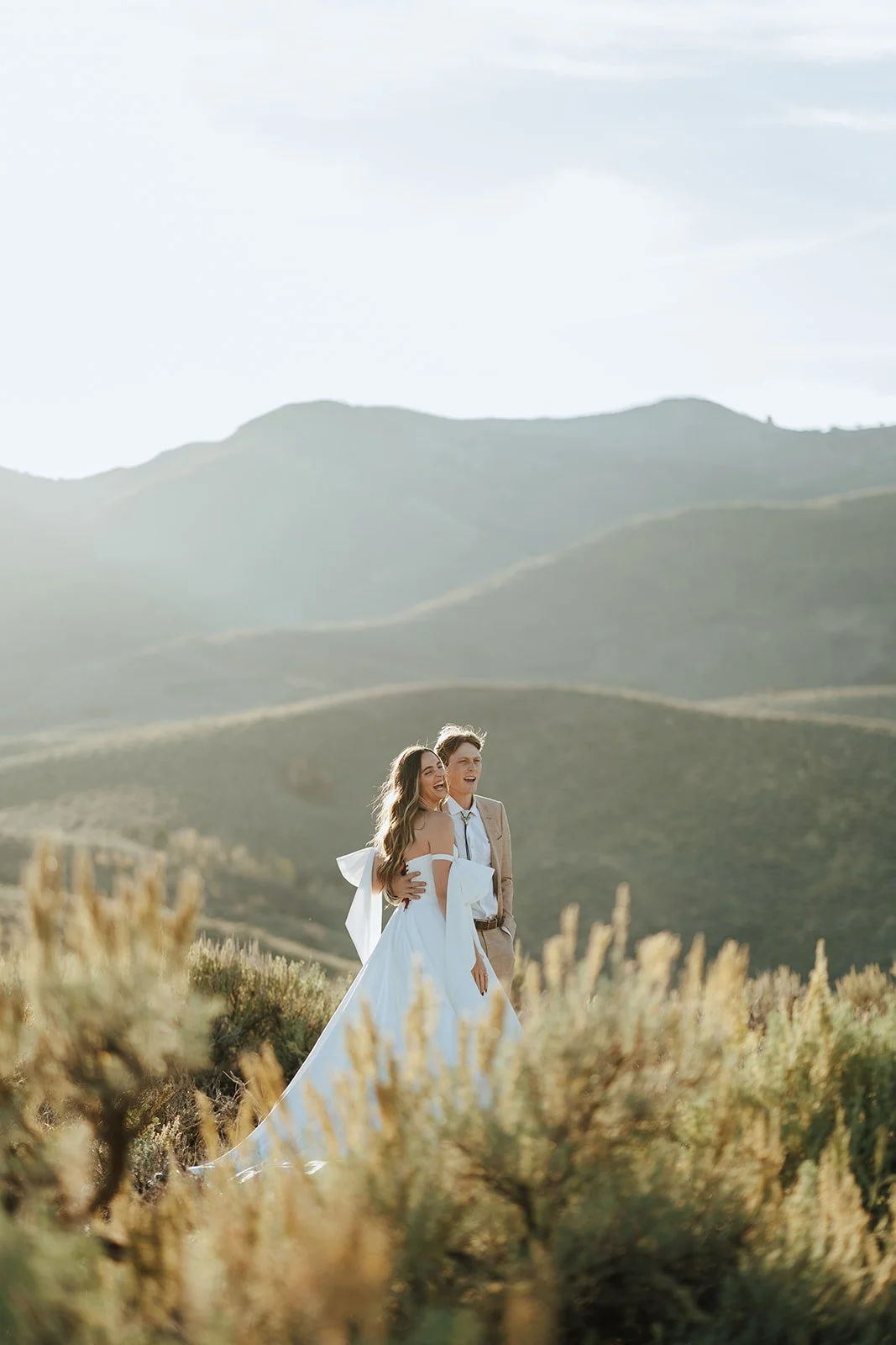 A bride and groom in a wedding gown and a tan suit stand in a meadow with mountainous peaks behind them - photography by Poise and Ivy Imagery