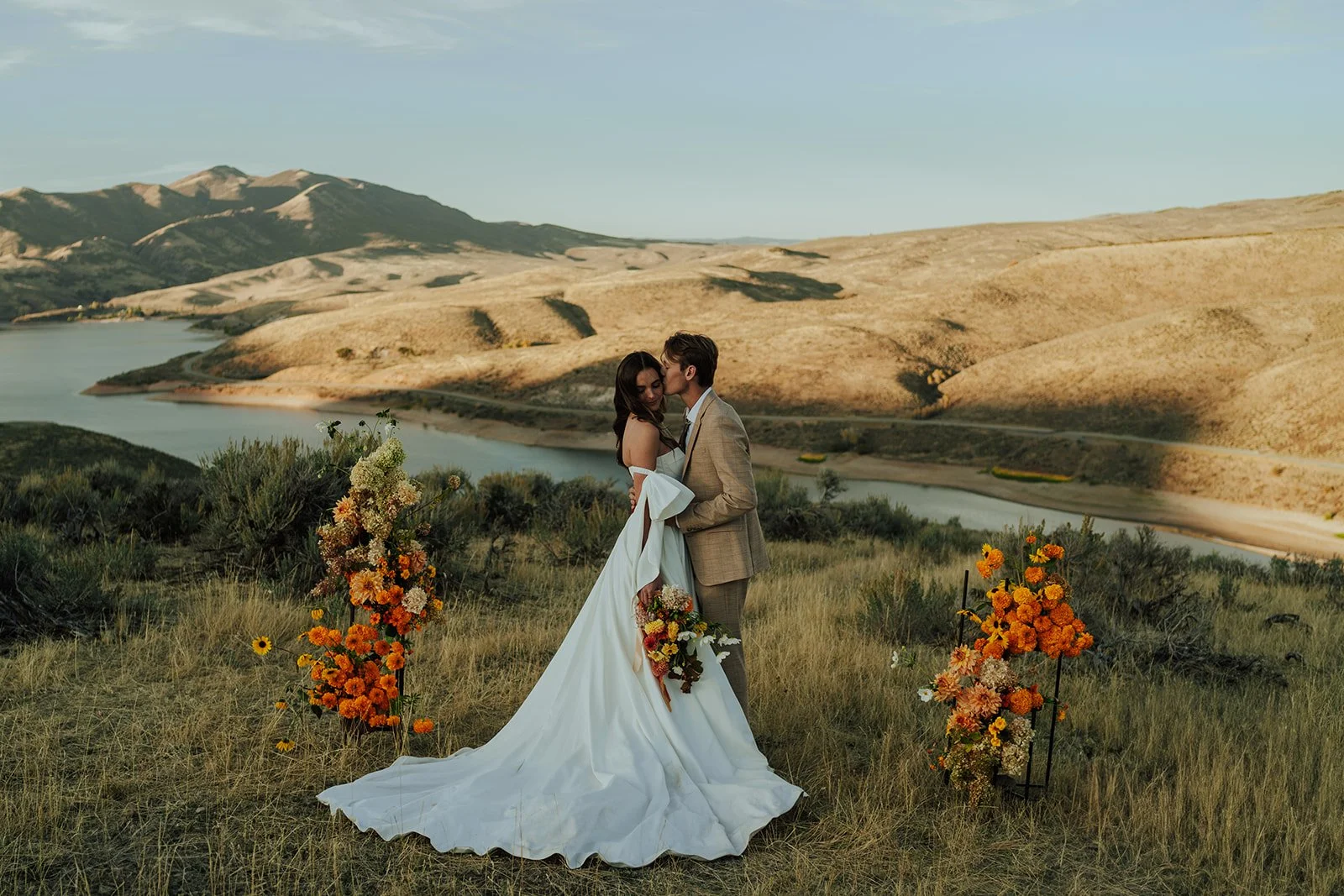 A bride and groom in a wedding gown and a tan suit stand in a meadow with mountains and a lake behind them - photography by Poise and Ivy Imagery