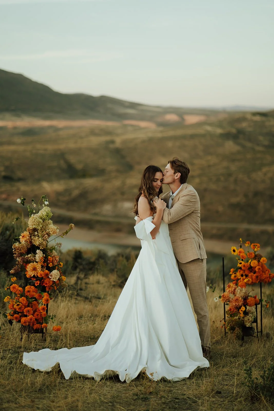 A bride and groom in a wedding gown and a tan suit stand in a meadow with mountains and a lake behind them - photography by Poise and Ivy Imagery