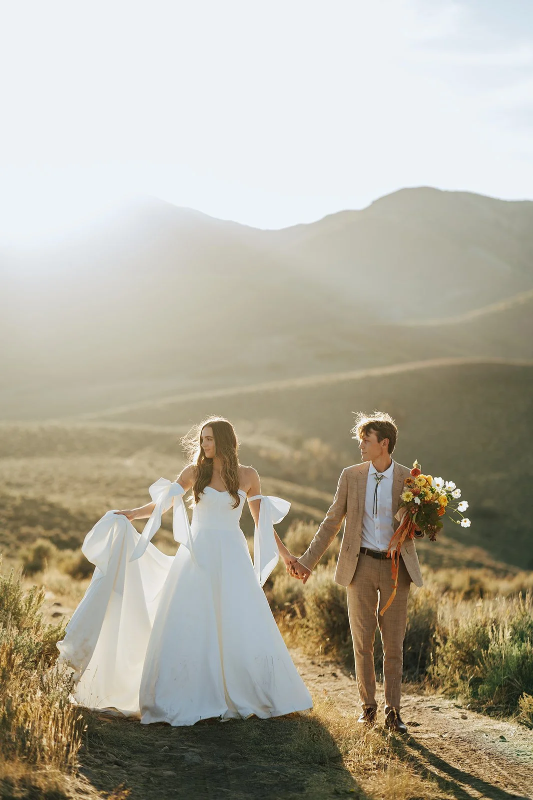 A bride and groom in a wedding gown and a tan suit stand in a meadow with mountainous peaks behind them - photography by Poise and Ivy Imagery