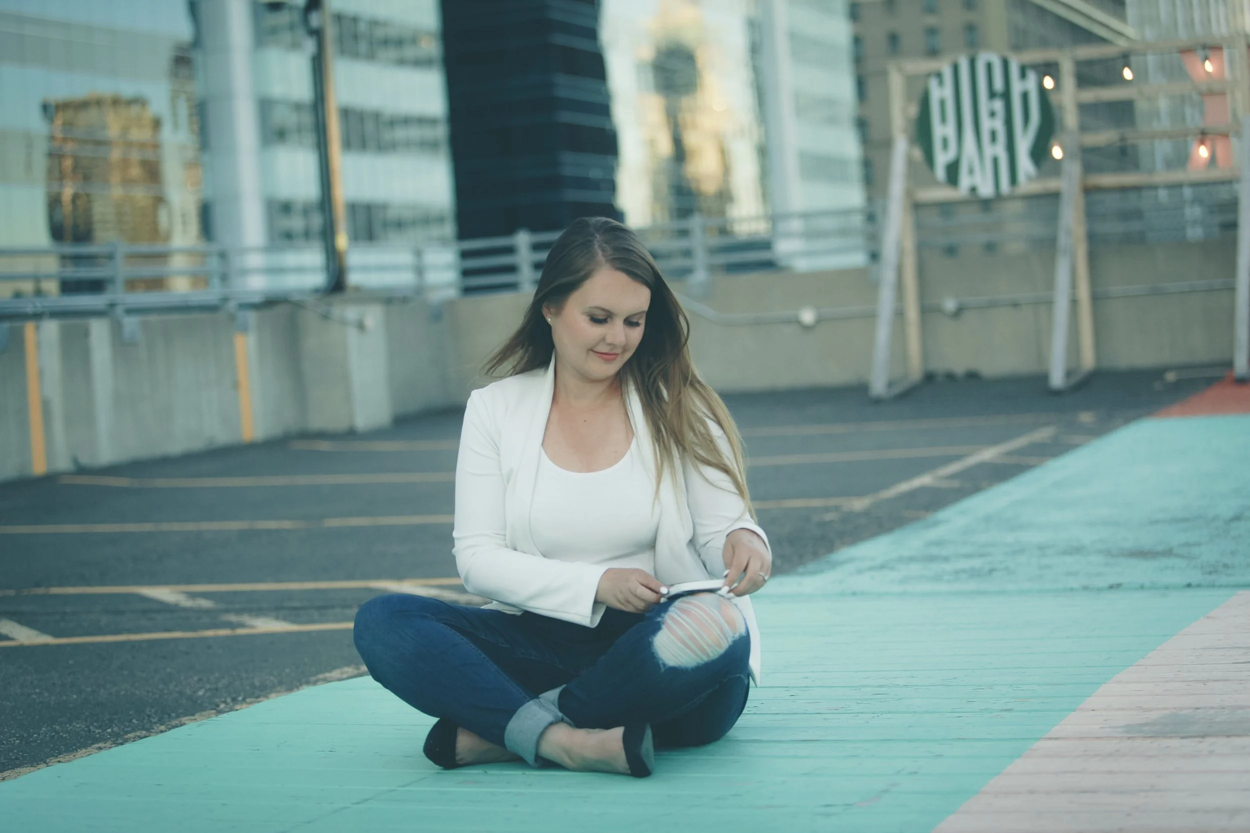 A woman with long hair, wearing a white blazer and ripped jeans, sitting cross-legged on a teal sidewalk in an urban area, using her phone.