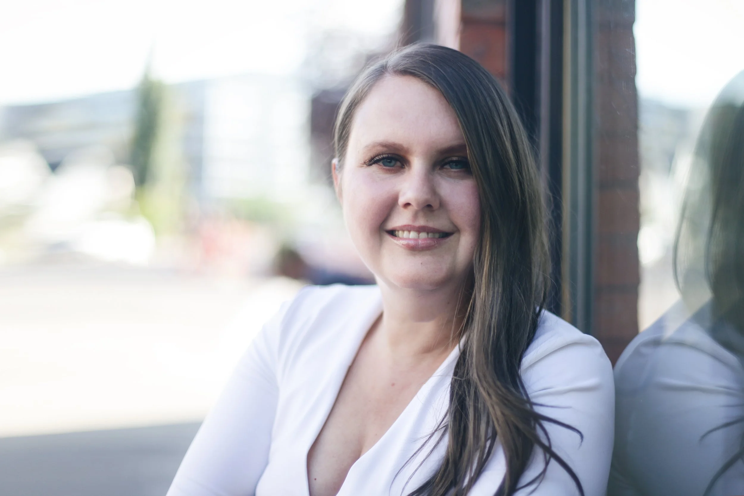 A woman with long dark hair and blue eyes smiling while sitting near a window.