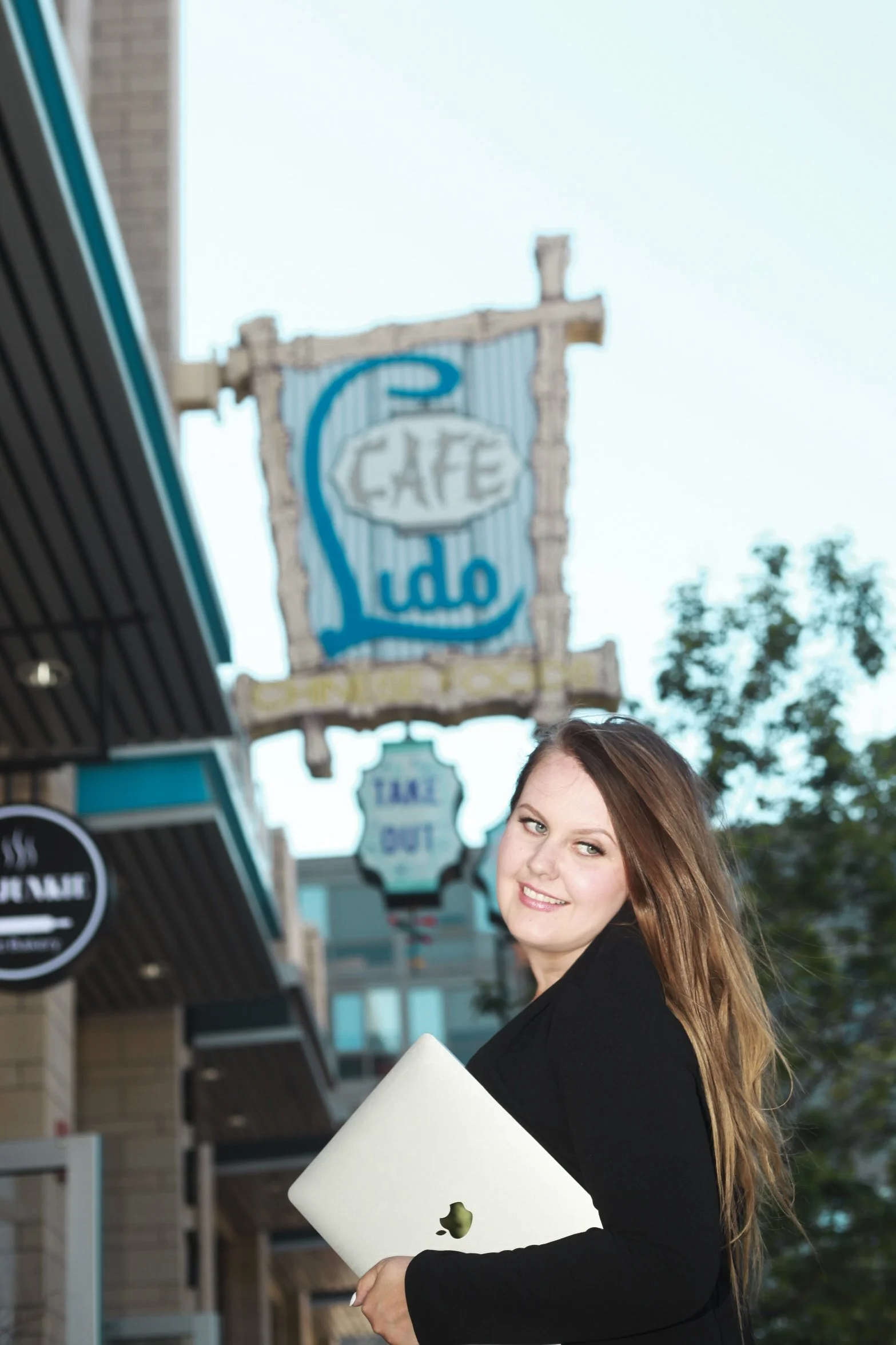 A woman with long brown hair holding a white MacBook laptop outside a cafe with a sign that says 'Cafe'.