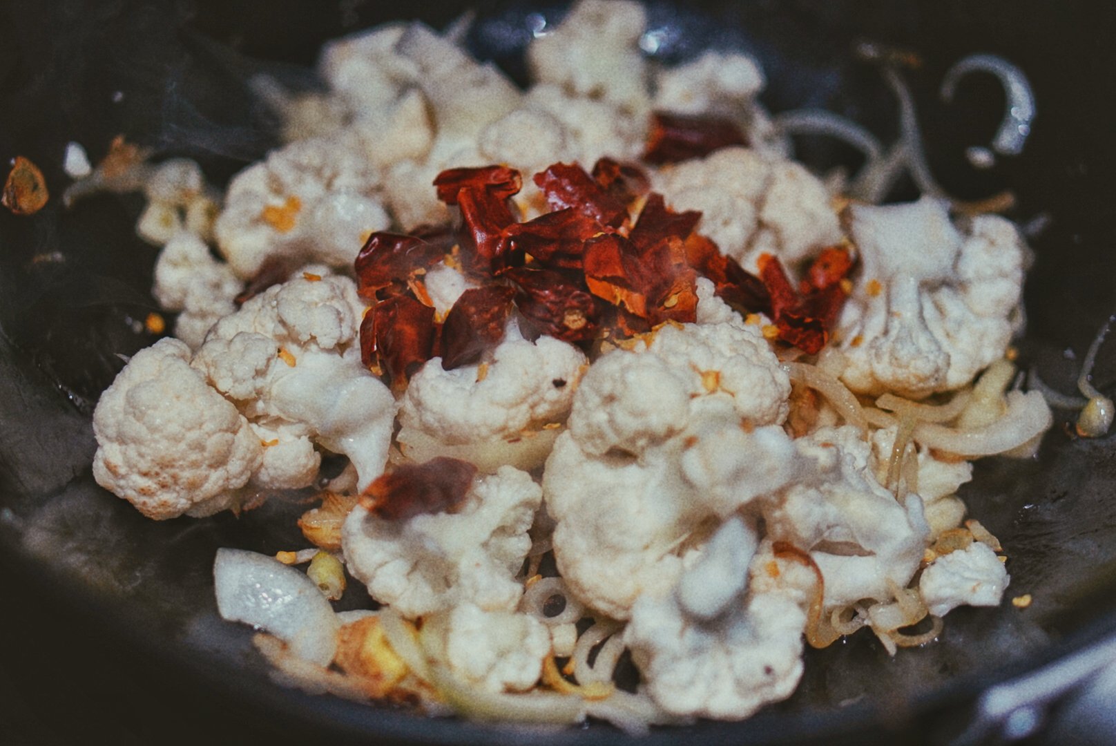 A close up shot of cauliflower, shallot, and dried chilis in a smoking hot cast aluminum wok on a stove. Shot and styled by Ellen Avigliano.