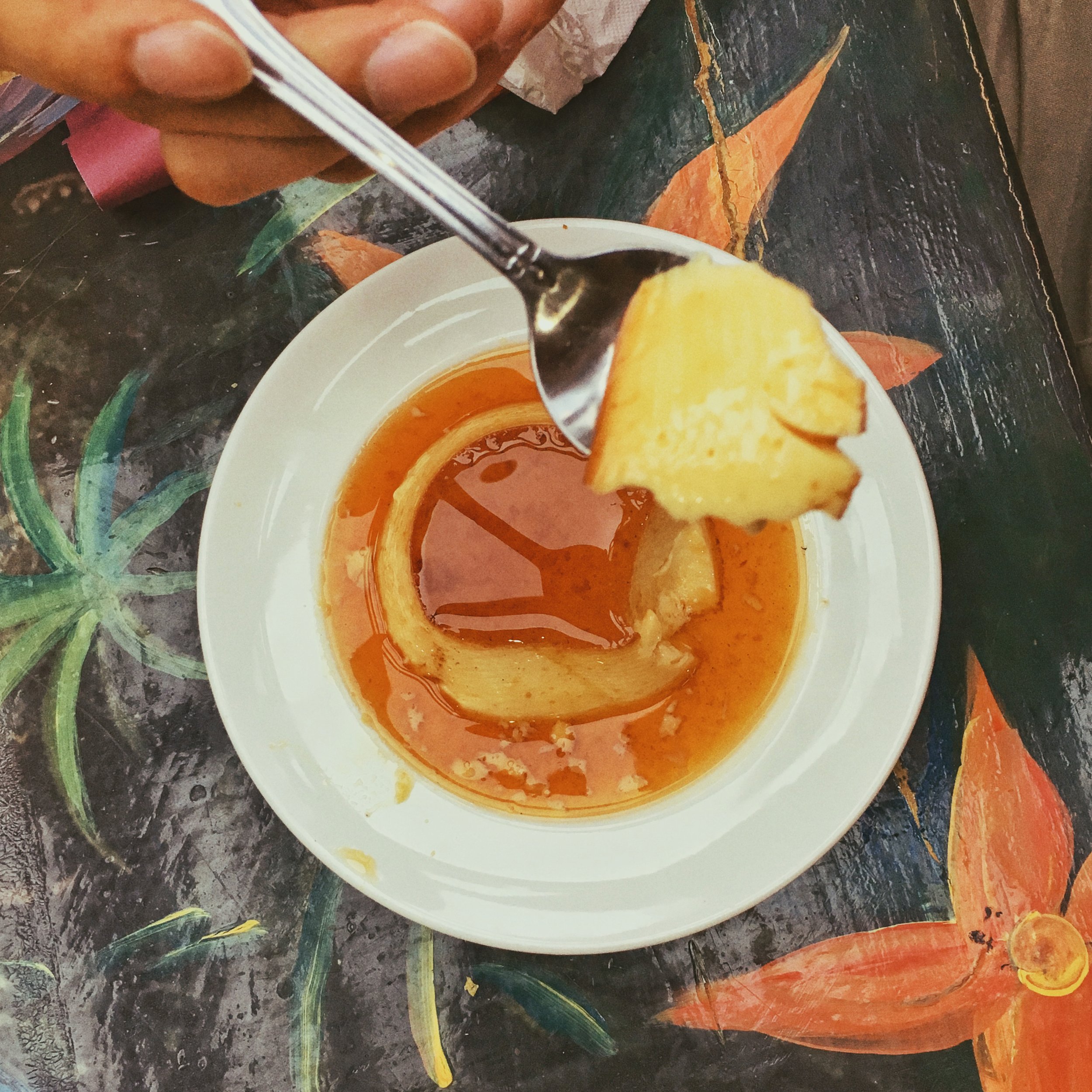 An overhead shot of a Cuban man eating a bite of flan at a restaurant against a backdrop of a tropical motif table, the spoon raised above the flan which is beautiful caramelized in a sauce. Food Photography shot by Ellen Avigliano.