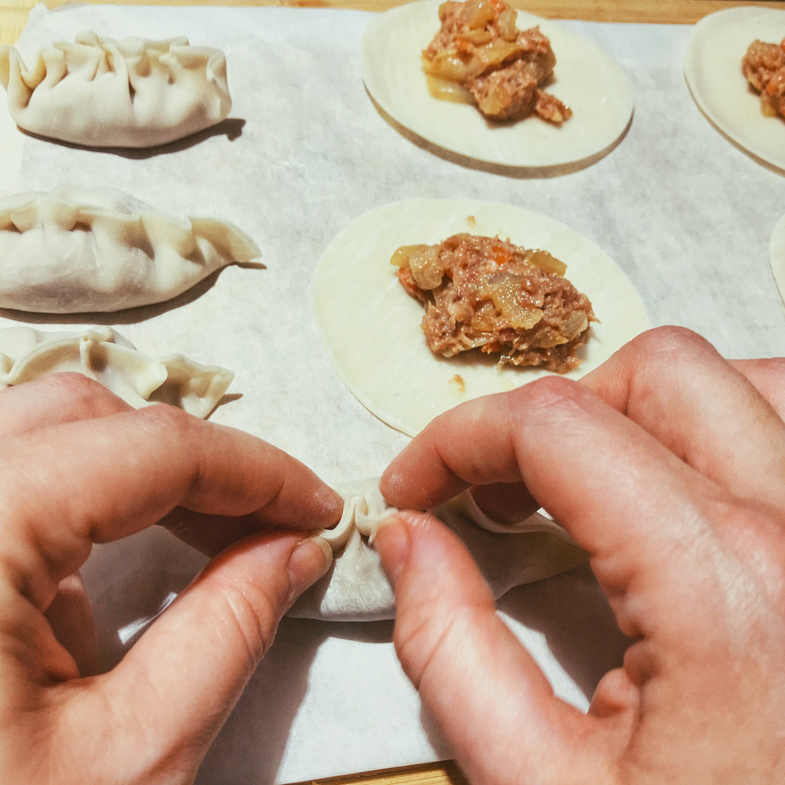 Handmade dumplings for Chinese new year arranged in a diagonal pattern in a monochromatic photo on a bamboo cutting board with parchment paper beneath it. Styled, cooked, assembled, and food photography shot by Ellen Avigliano.