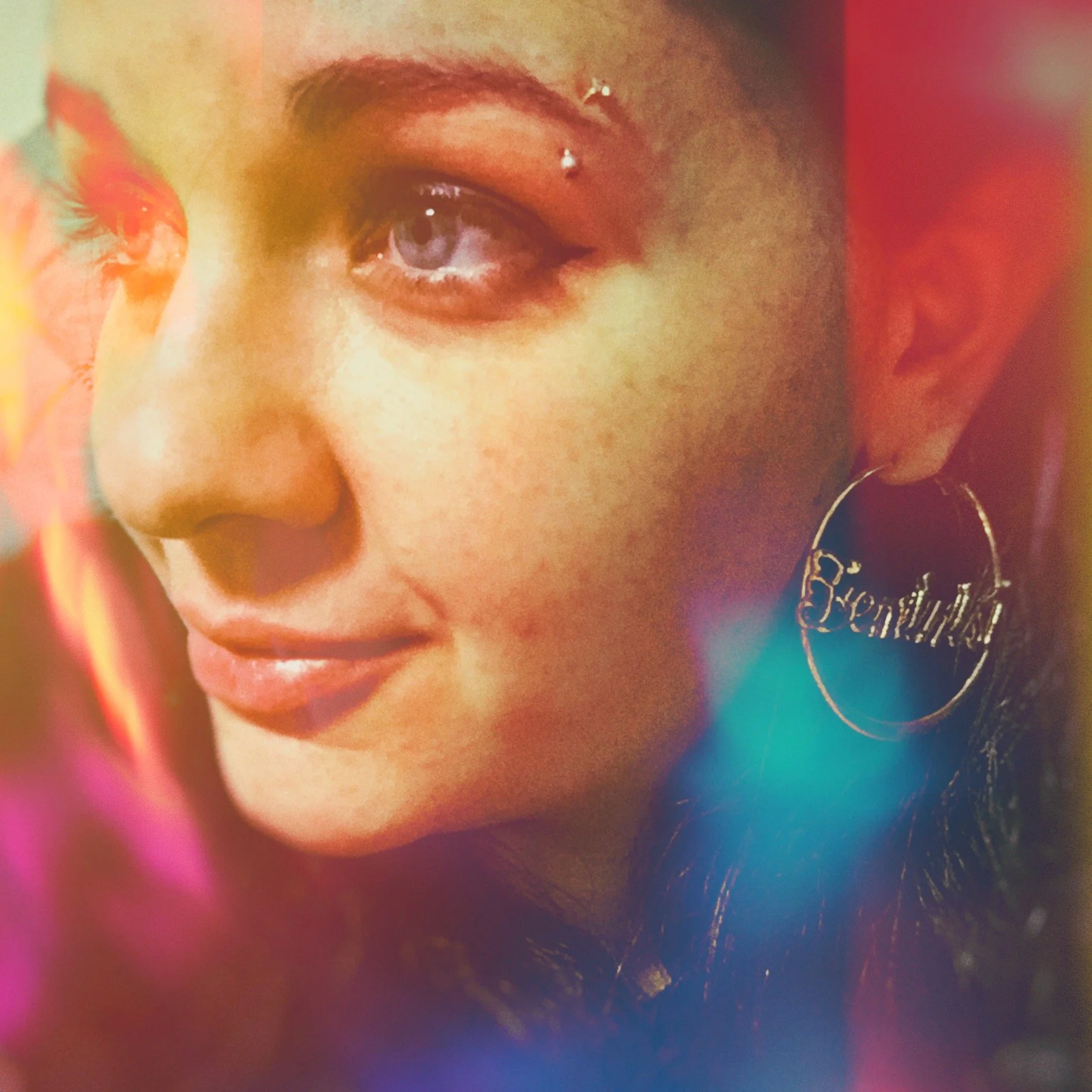 Close-up of a woman's face, showing her eye, nose, and lips, with a colorful, blurry background and a large earring with the word 'Beautiful'.