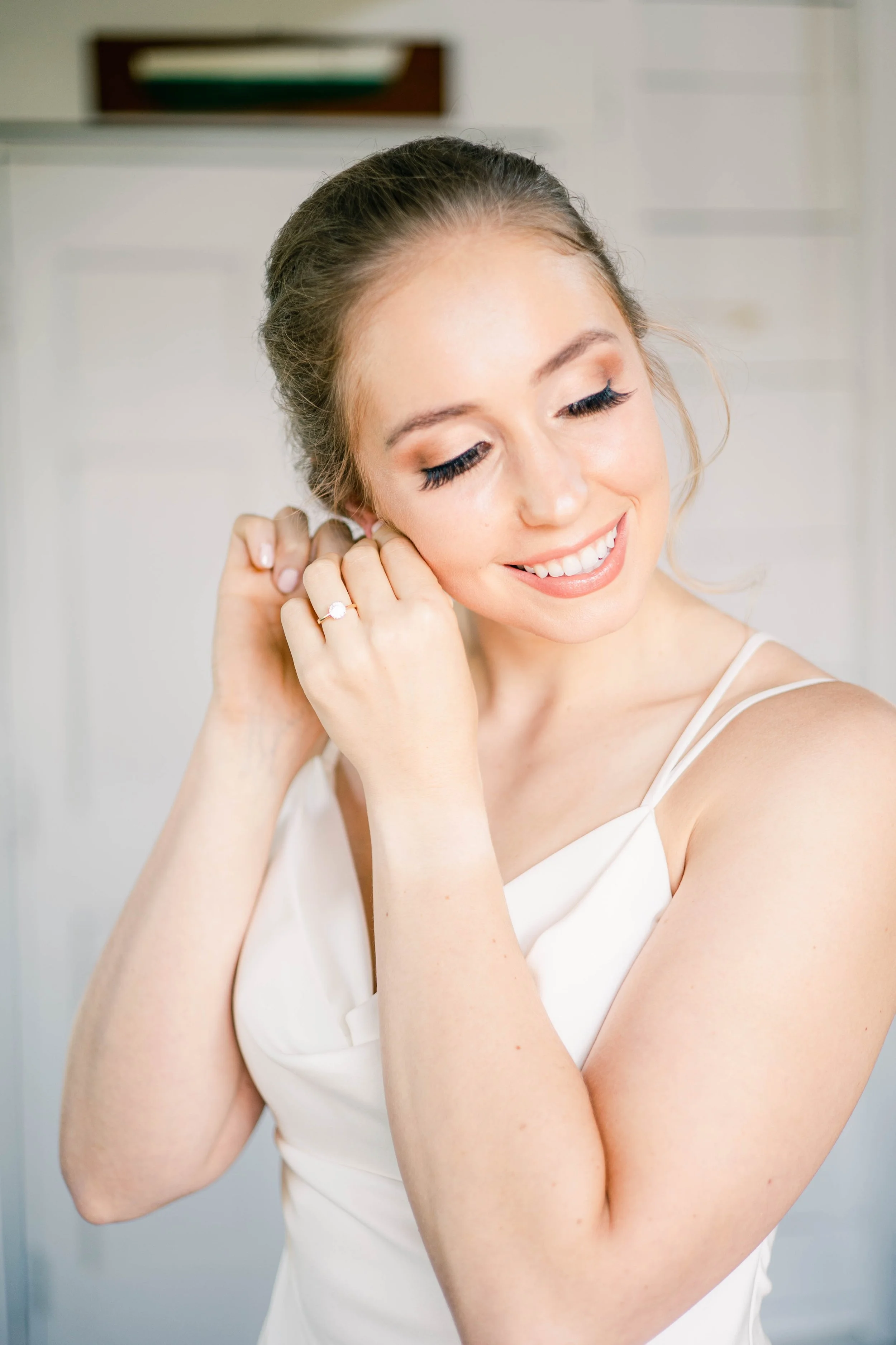 A young woman with fair skin and light brown hair styled in an updo is putting on earrings. She has makeup with neutral tones and is smiling with her eyes closed. She is wearing a white satin dress with thin straps.