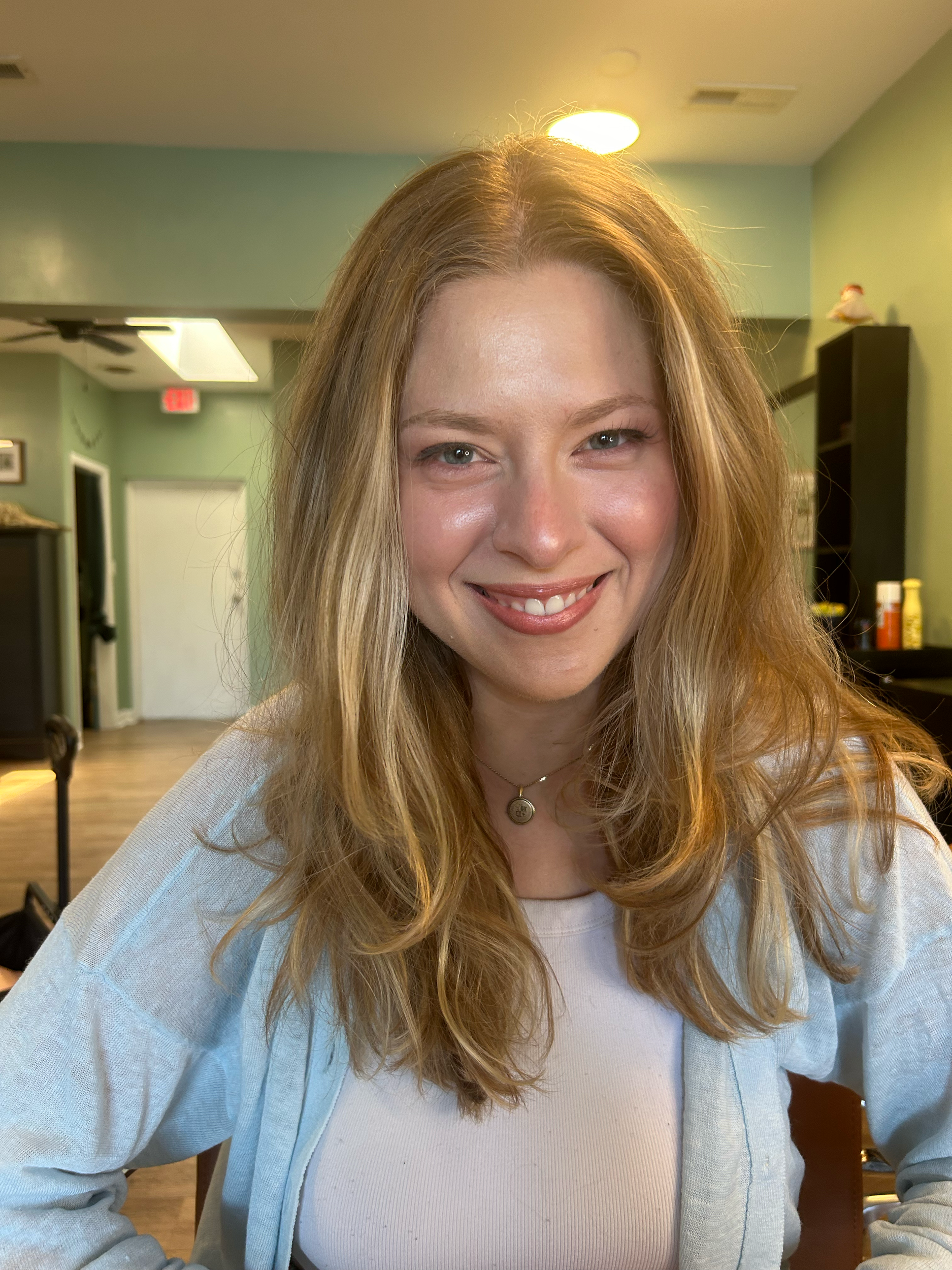 A woman with long, wavy, blonde hair smiling in a room with green walls and black shelves.