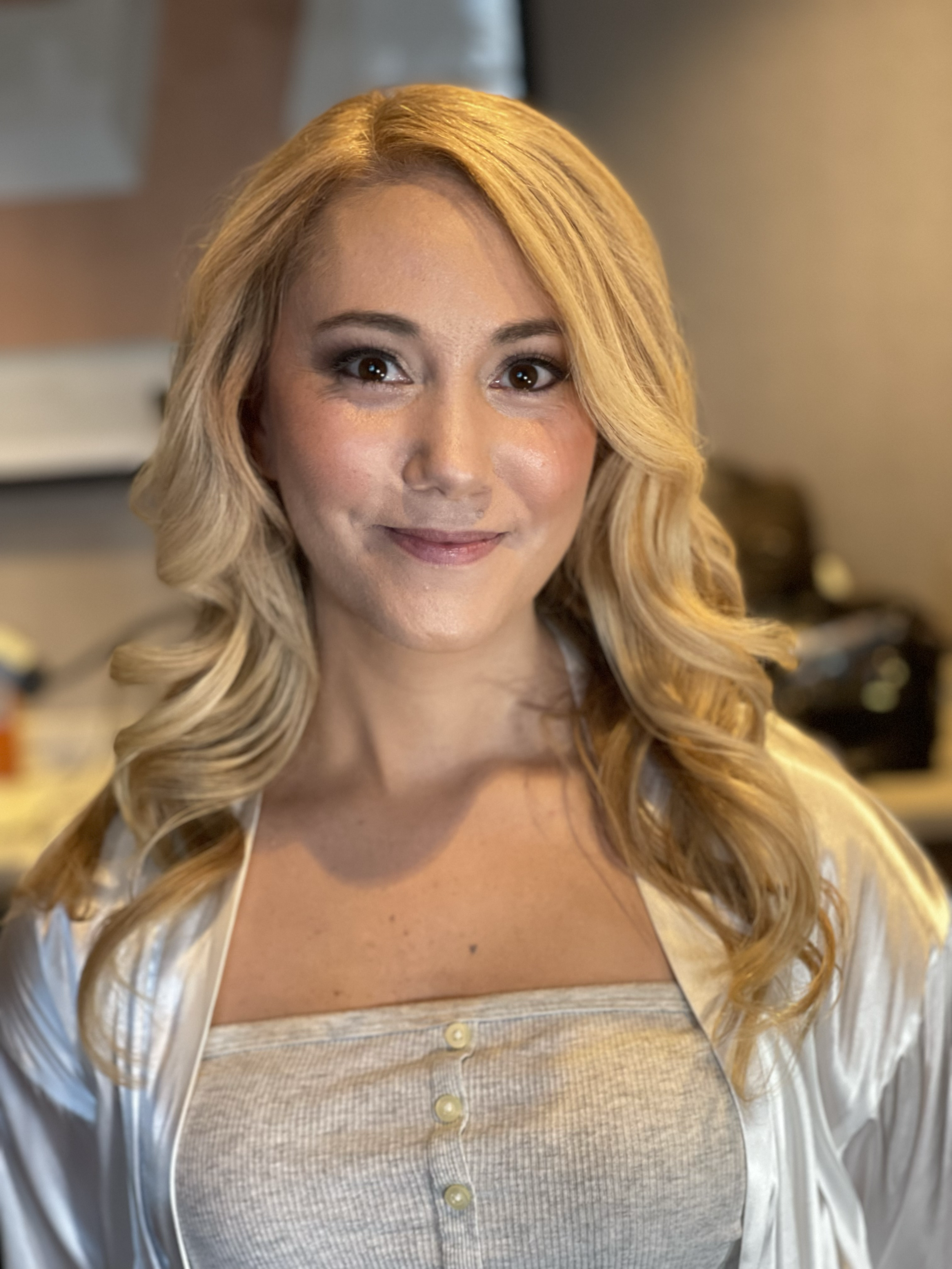 A woman with long blonde hair, wearing a beige top and satin robe, smiling indoors.