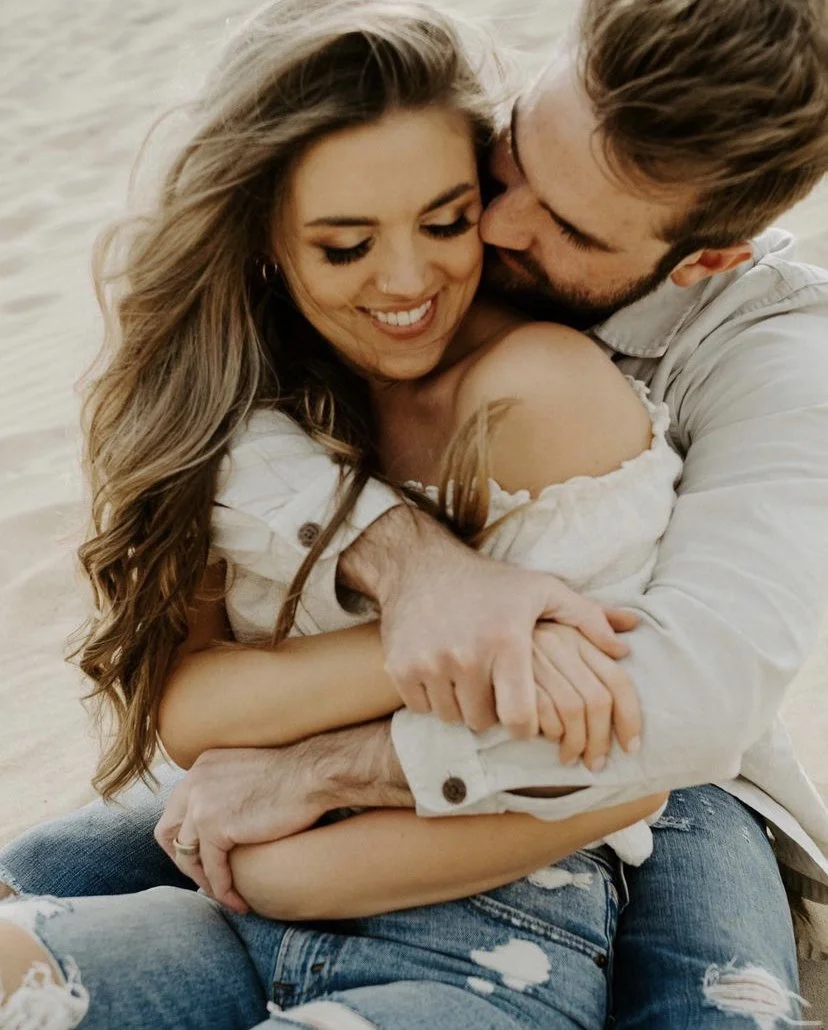 A loving couple embracing and smiling on a beach, with the man kissing the woman on the cheek.