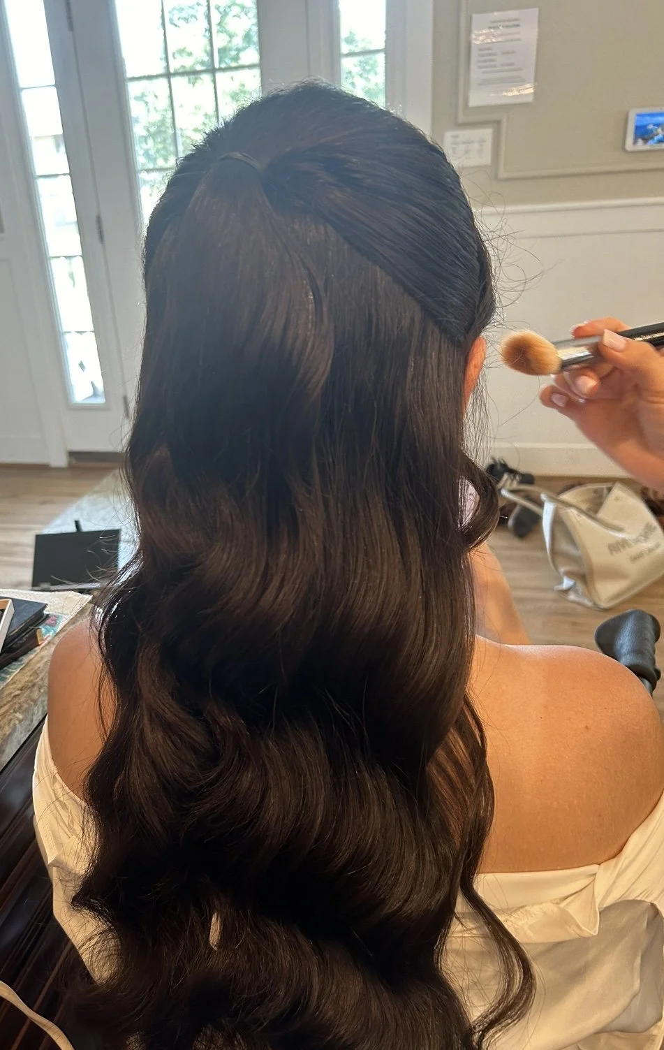 A woman with long, wavy dark hair is getting her hair styled with a curling iron in a salon. A stylist is holding a makeup brush nearby.
