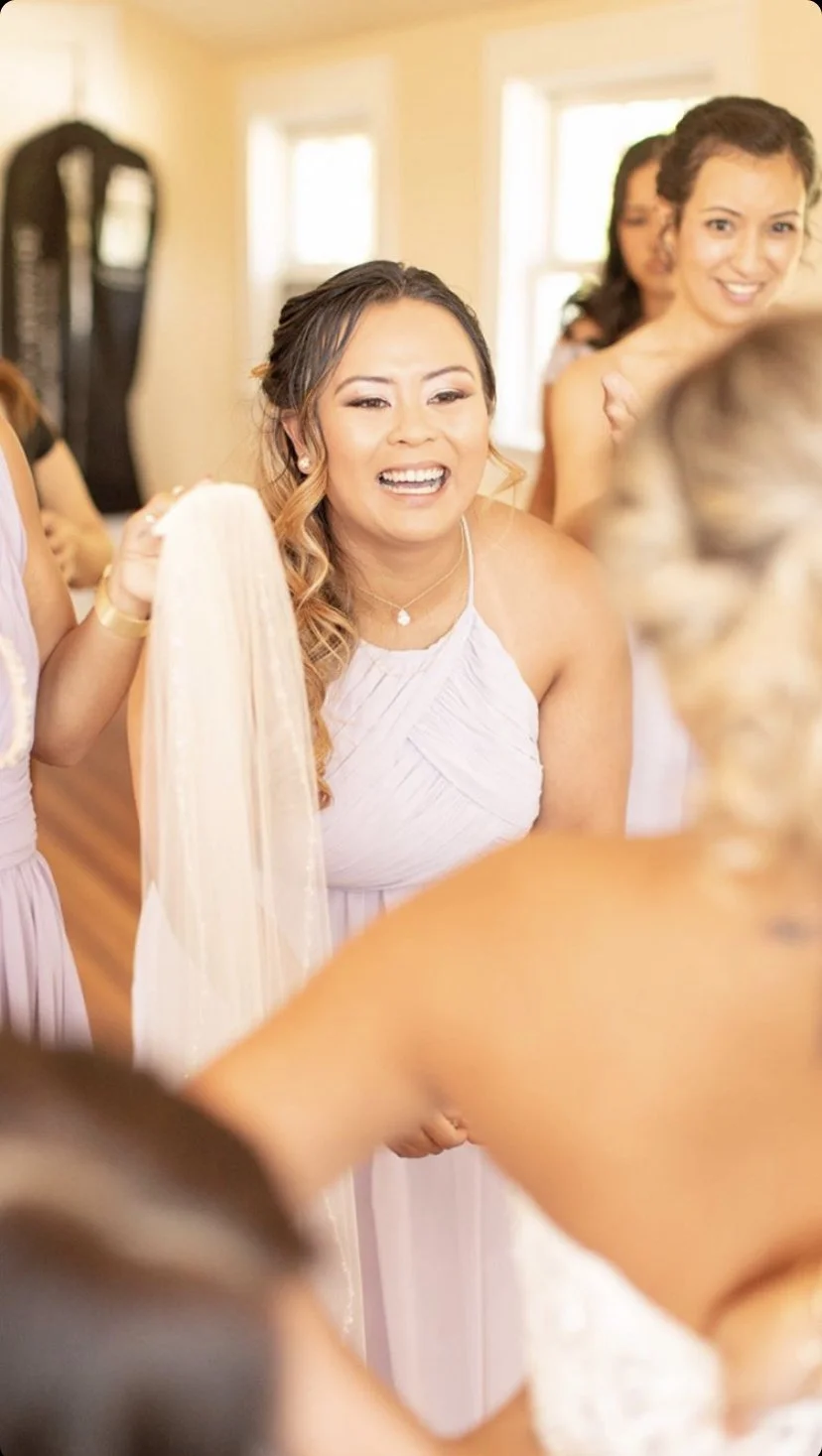 Women at a wedding event with one woman smiling and holding what appears to be a wedding dress or veil. The setting is bright with natural light coming from windows.