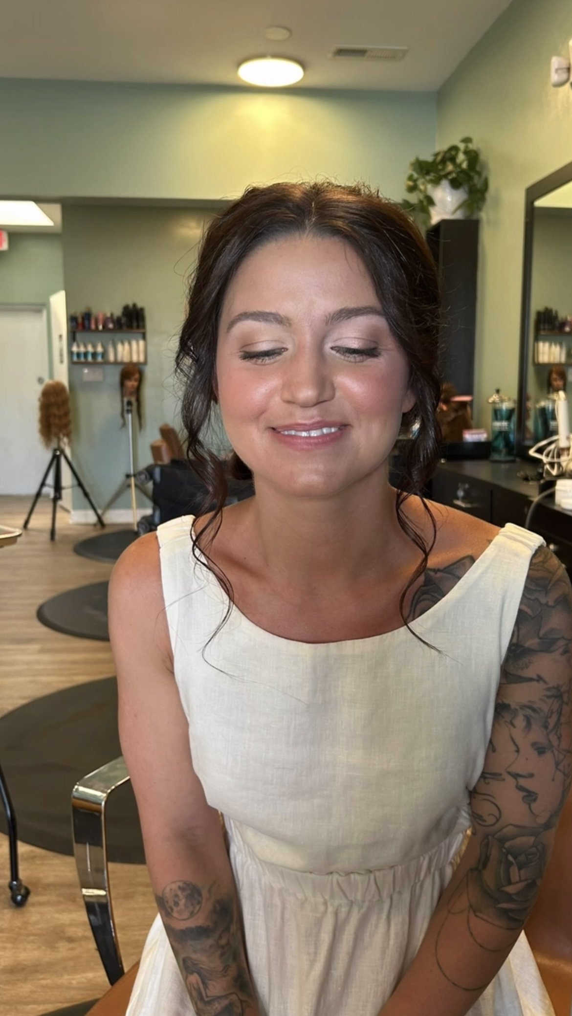 bride with glowing skin and rosy cheeks looking down, her hair is in an updo, she is wearing a white dress and there is a hair salon behind her