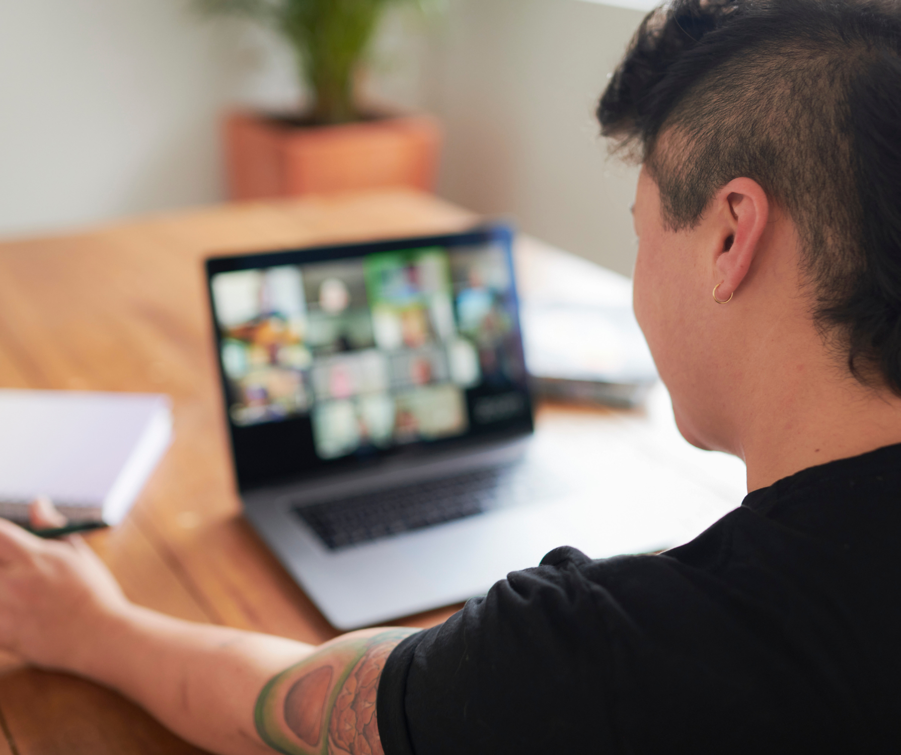 Person with a tattoo on their arm participating in a virtual meeting on a laptop, visible on the screen are multiple video call windows, sitting at a wooden desk with papers and a notebook, in a room with a plant in the background.