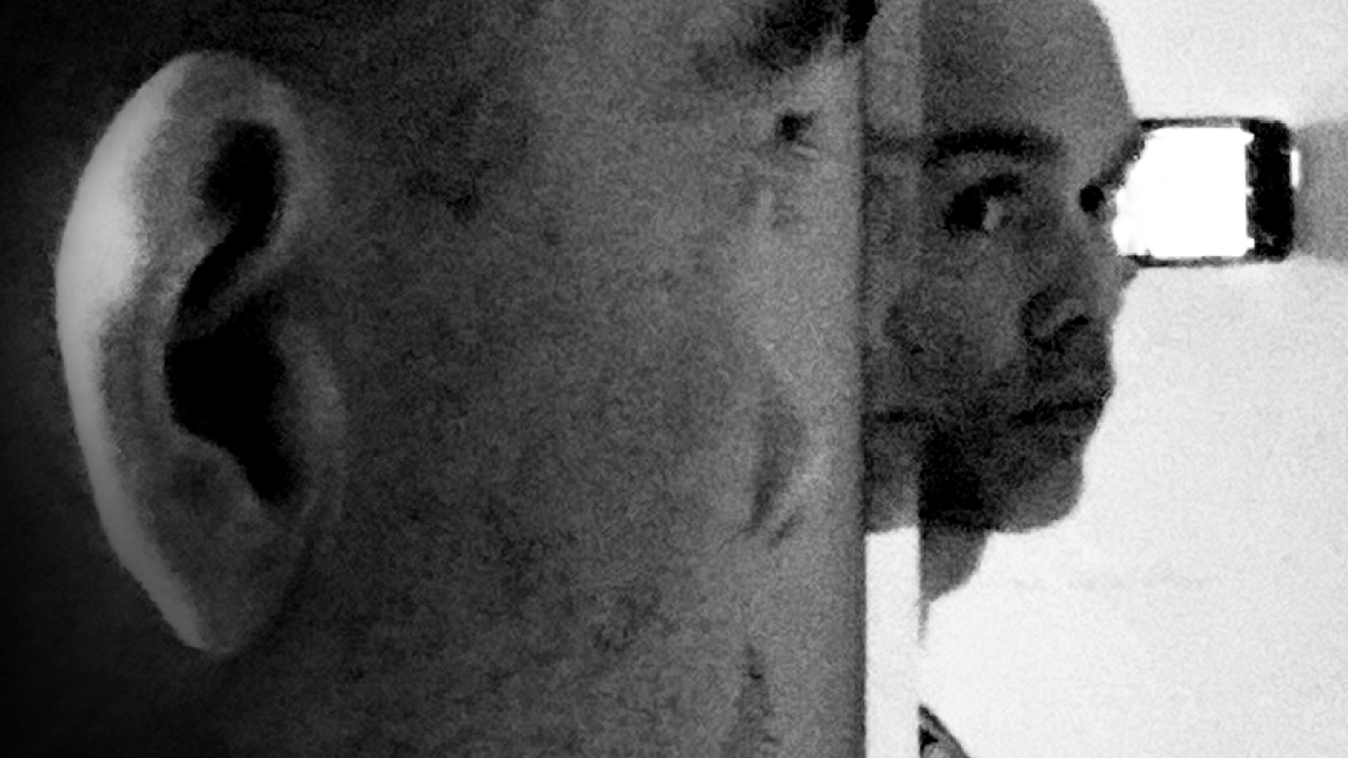 Close-up black-and-white photo of a man taking a photograph, with his head turned to the side, in front of a mirror showing his reflection in such a way that there appears to be more than one person.