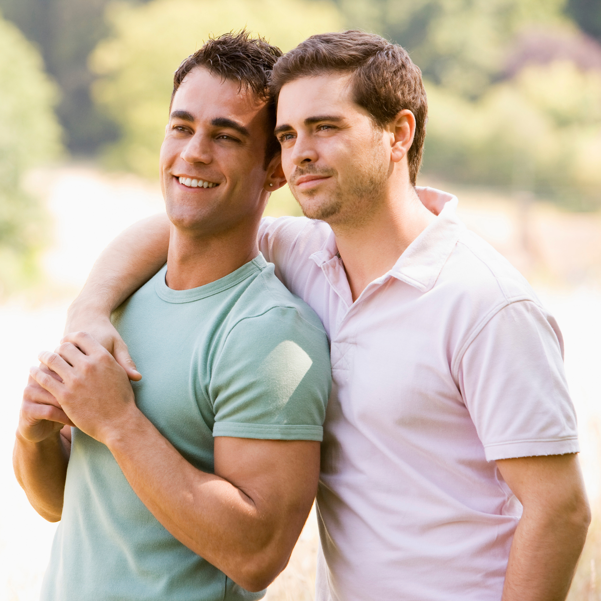 Two men stand outdoors, smiling, with one arm around each other's shoulders, enjoying a sunny day in a park or nature setting.