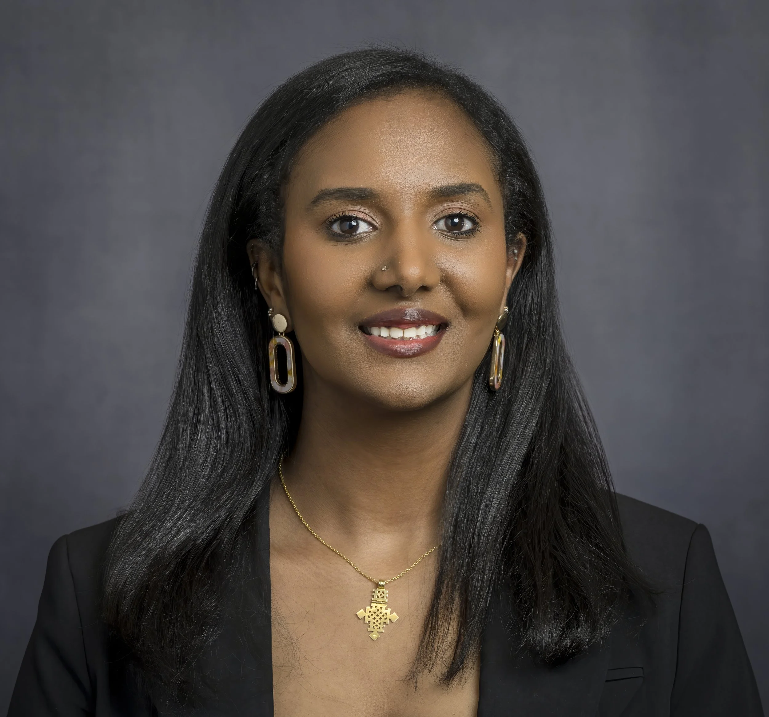 A professional woman with long black hair, wearing earrings, a necklace, and a black blazer, smiling at the camera against a gray background.