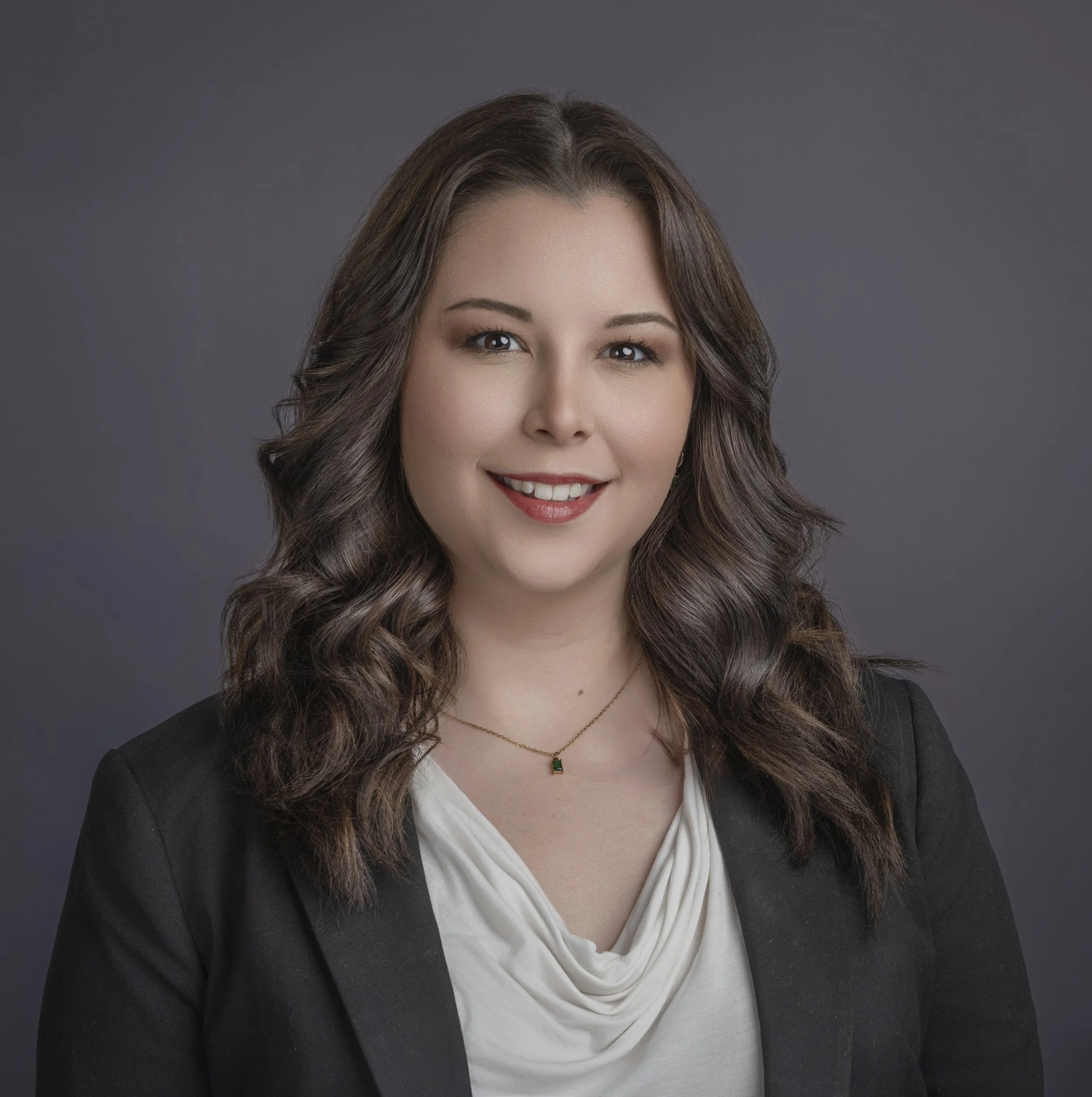 Professional headshot of a woman with wavy brown hair, wearing a black blazer and white top, smiling against a gray background.
