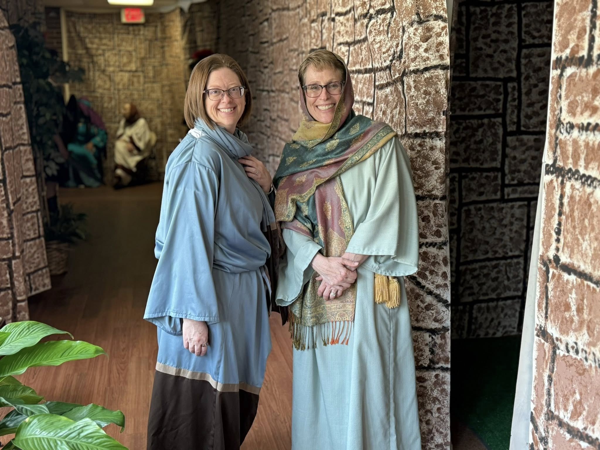 Two Journey to Easter volunteers in blue attire smiling in front of painted backdrops.
