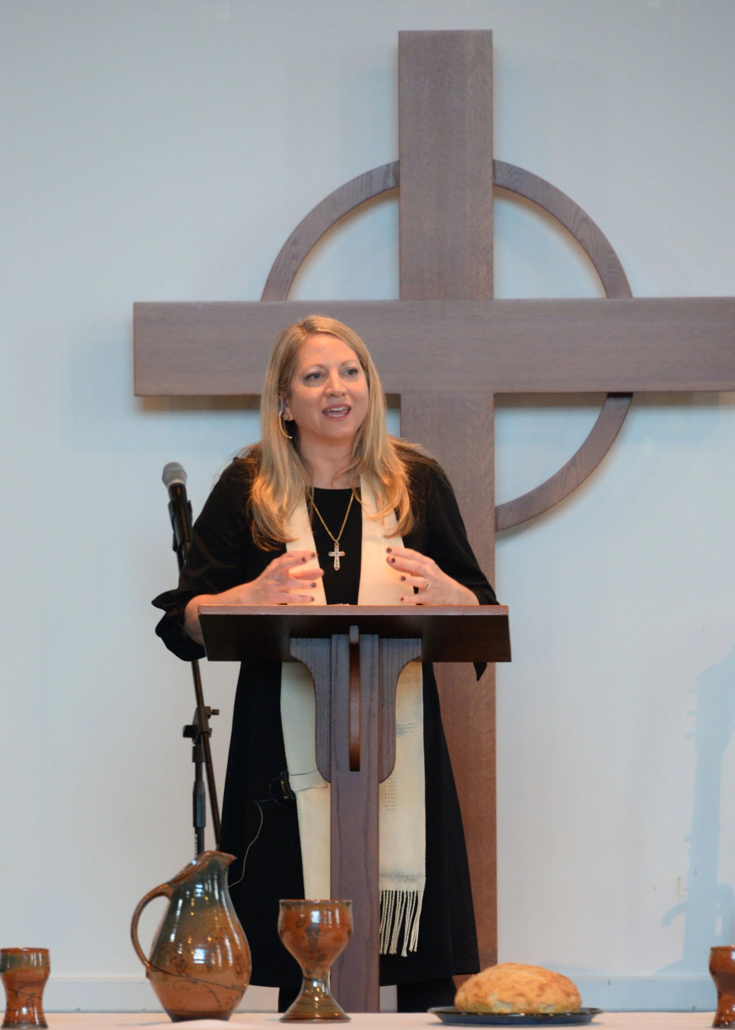 A woman standing at a lectern giving a speech, with a large wooden cross in the background, on a wall behind her, and a table with ceramic pitchers and a loaf of bread in the foreground.