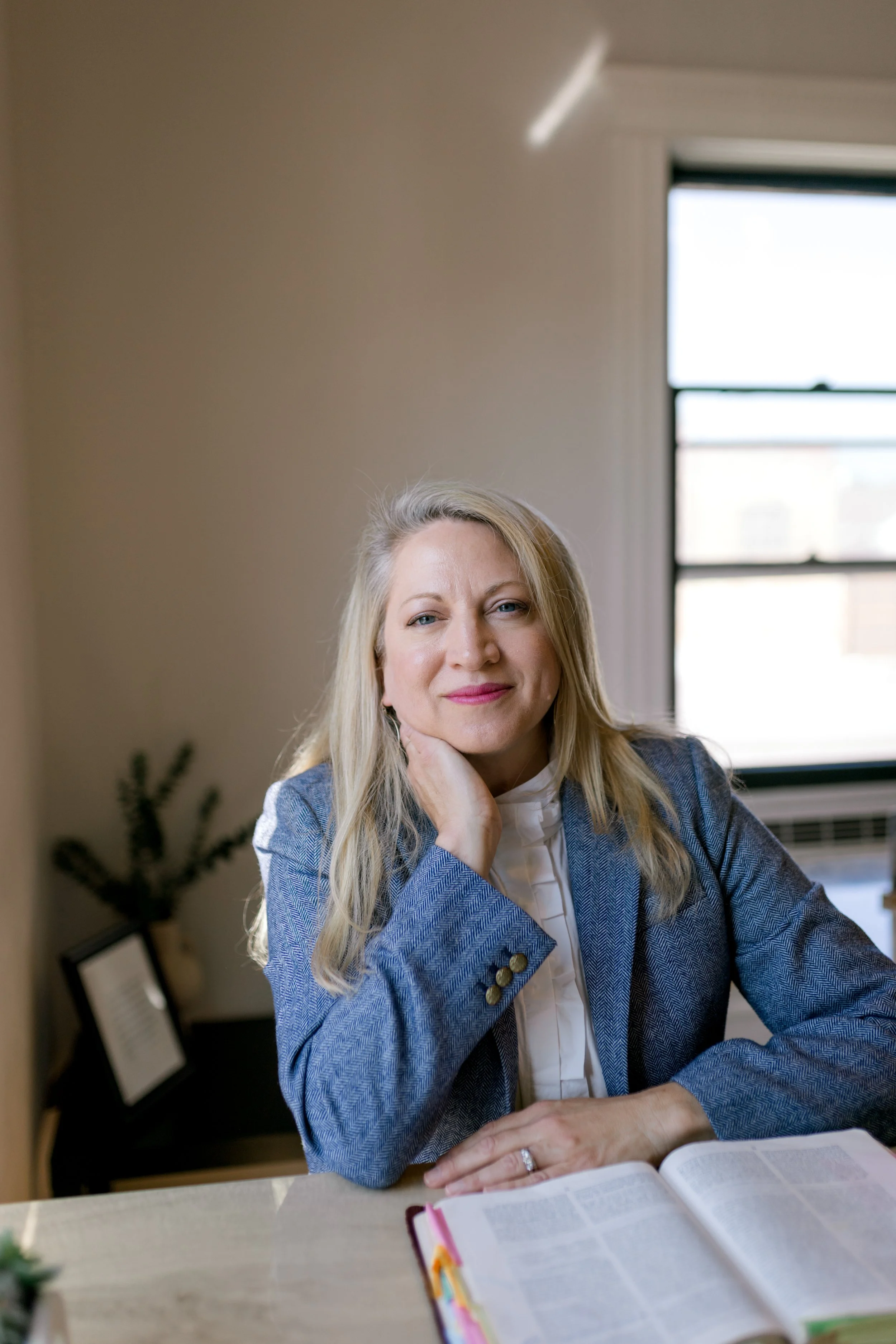 A woman with blonde hair, wearing a gray blazer and white blouse, sitting at a desk with an open book or planner, resting her chin on her hand, in a well-lit room with a window in the background.