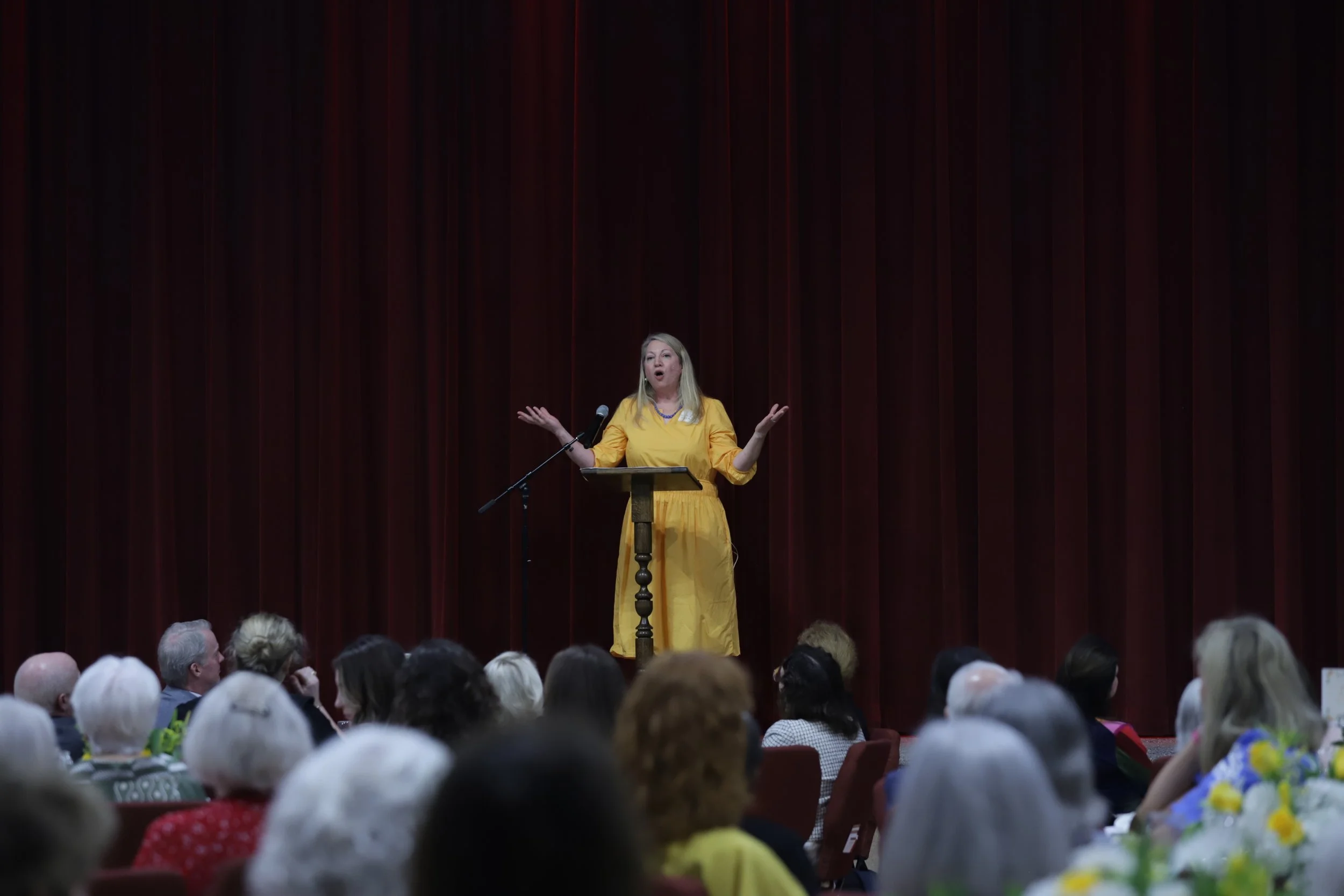 A woman in a yellow dress speaking at a podium on a stage with a dark red curtain backdrop, addressing an audience of older adults.