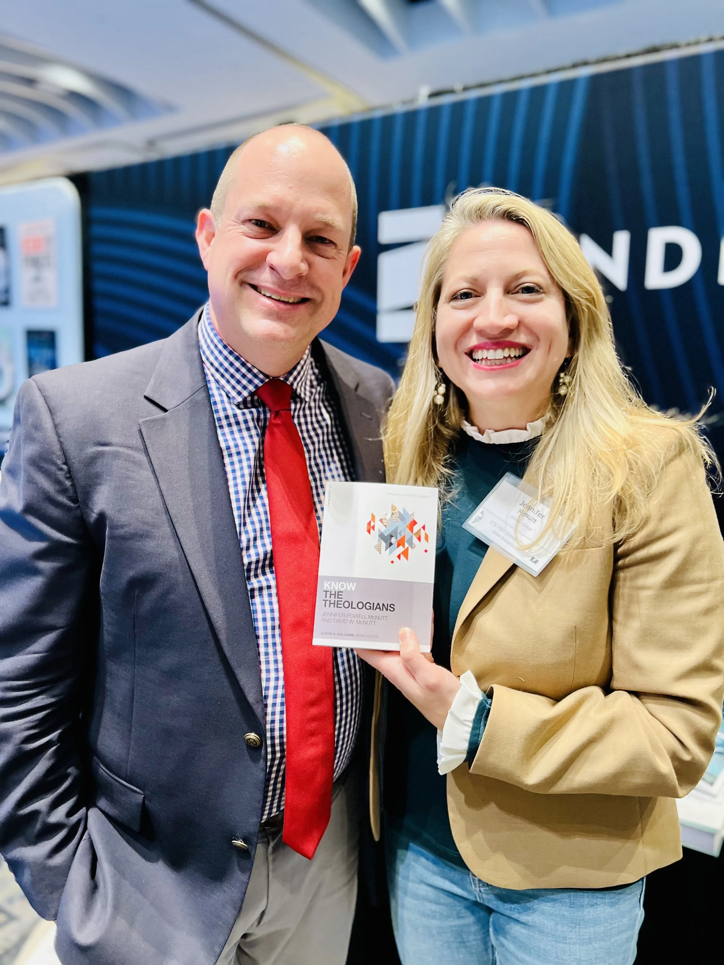 Two people smiling at a professional event, one holding a book titled 'Know the Theologians' and wearing a name badge, with a blue backdrop and vending machines in the background.