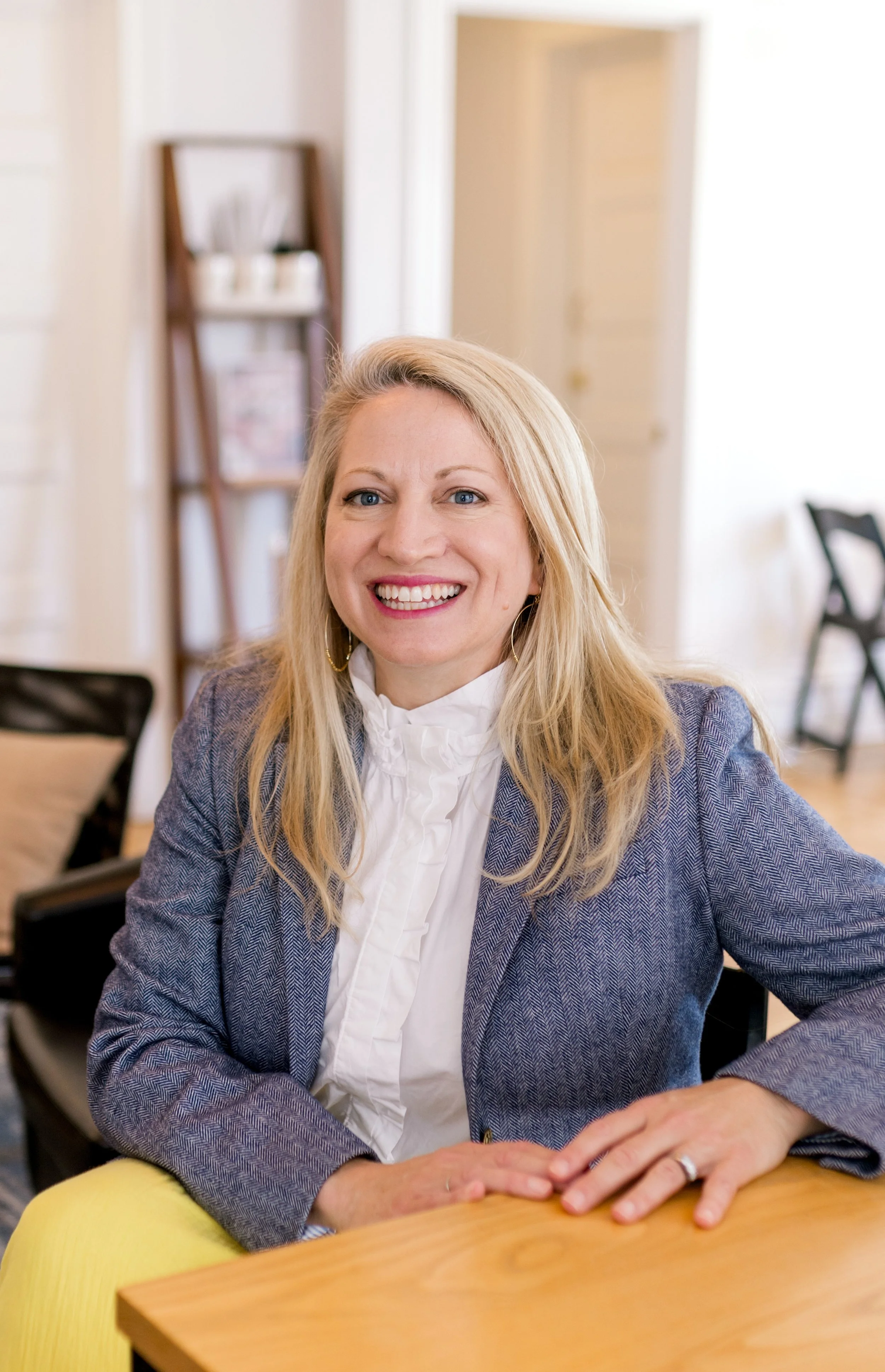 A woman with blonde hair smiling, wearing a blue blazer and white ruffled blouse, sitting at a wooden table in a bright room with white walls, a bookshelf in the background, and chairs around.