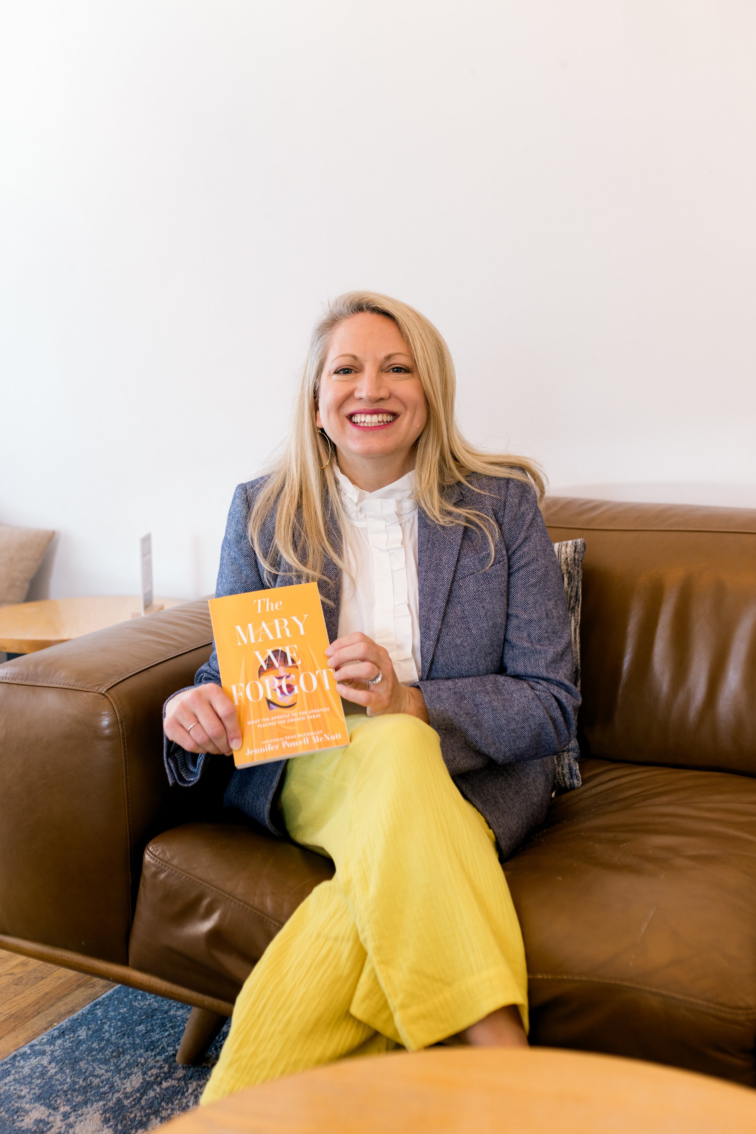 A woman with blonde hair is sitting on a brown leather couch, smiling and holding a yellow book titled 'The MARY We FORGOT'. She is wearing a gray blazer, white shirt, and yellow pants, with a white wall in the background.