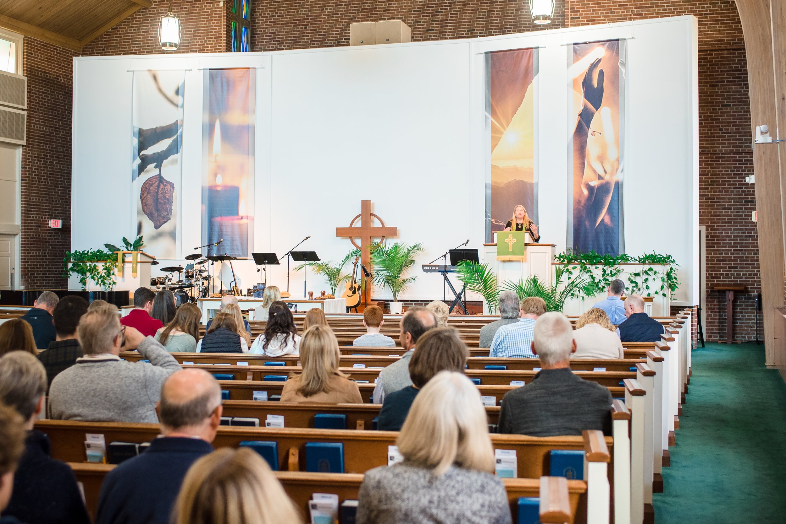 People gathered in a church listening to a woman speaking at the pulpit, with musical instruments on stage, a wooden cross, and large banners showing candles and hands in prayer or worship. The church has brick walls, high ceilings, and wooden pews.