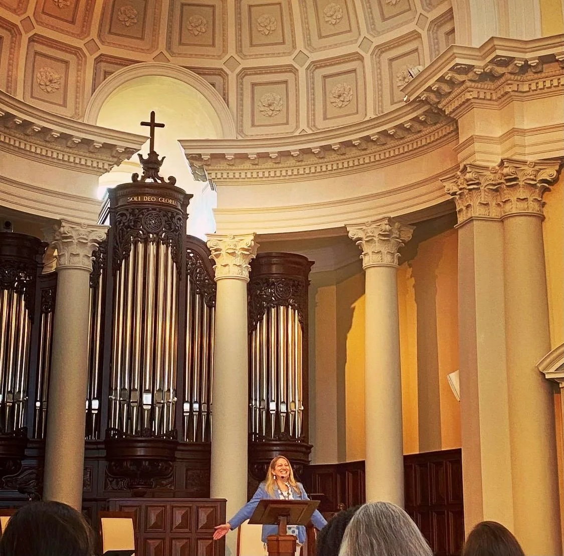 A woman standing at a podium smiling, with a large pipe organ behind her, in a church or cathedral with columns and a domed ceiling.