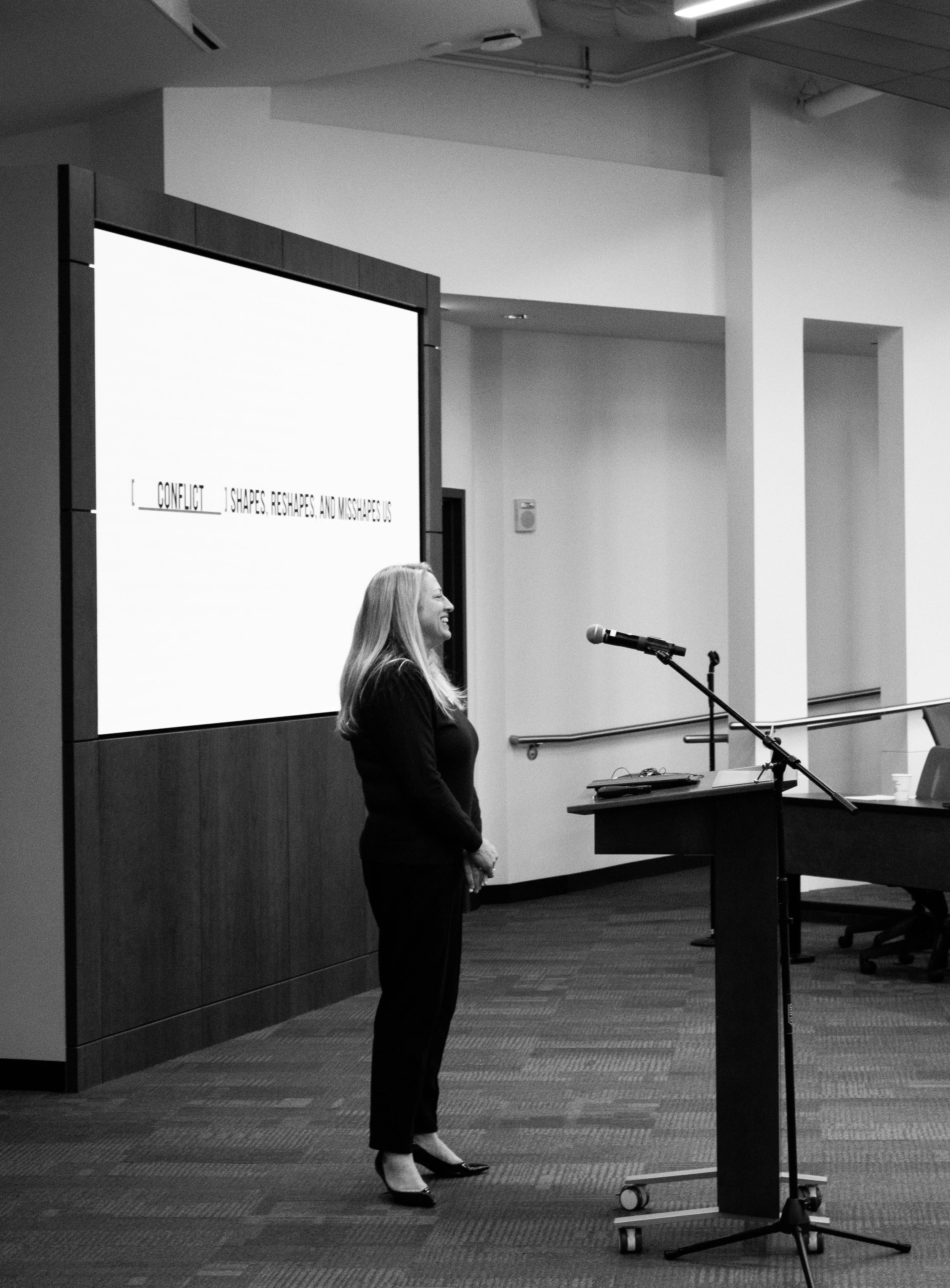A woman stands in front of a large screen displaying text, speaking into a microphone, in a modern conference room with a podium and a coffee cup on the table.