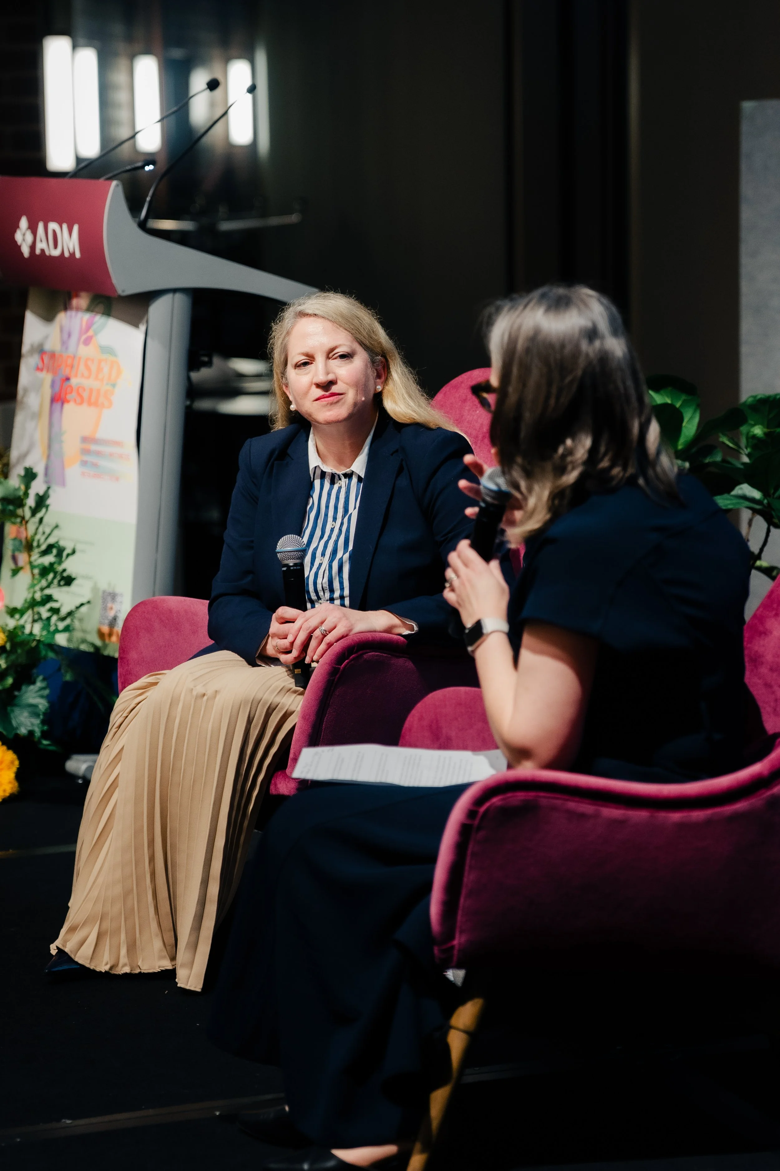 A woman with blonde hair, wearing a dark blazer and striped shirt, sitting on a pink chair holding a microphone, participating in a discussion at an indoor event with another woman, with a podium and banner that reads 'STEPPOSED JESUS' in the backgro