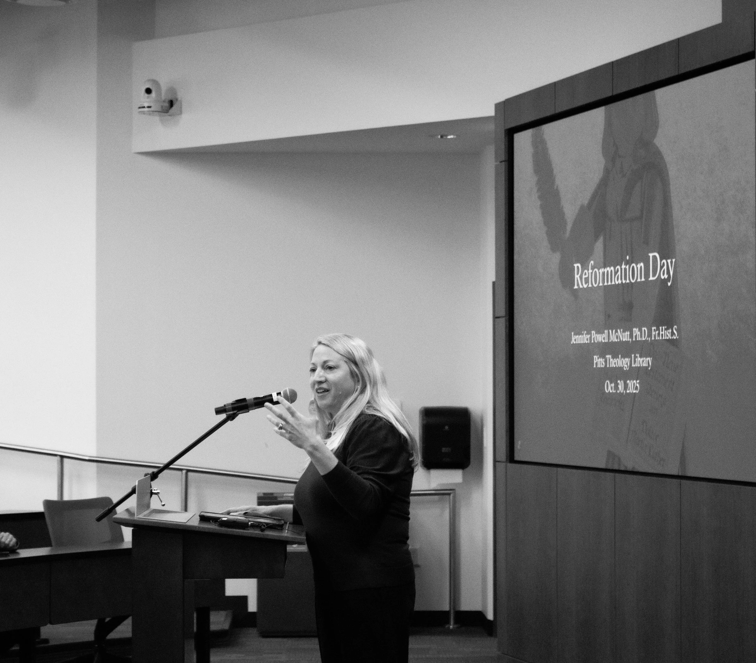 A woman standing at a podium, speaking into a microphone during a presentation or event. She has long, light-colored hair and is wearing a dark top. In the background, a large screen displays part of the presentation with the title "Reformation Day" and the name "Jennifer Powell McNutt, Ph.D." along with other text.