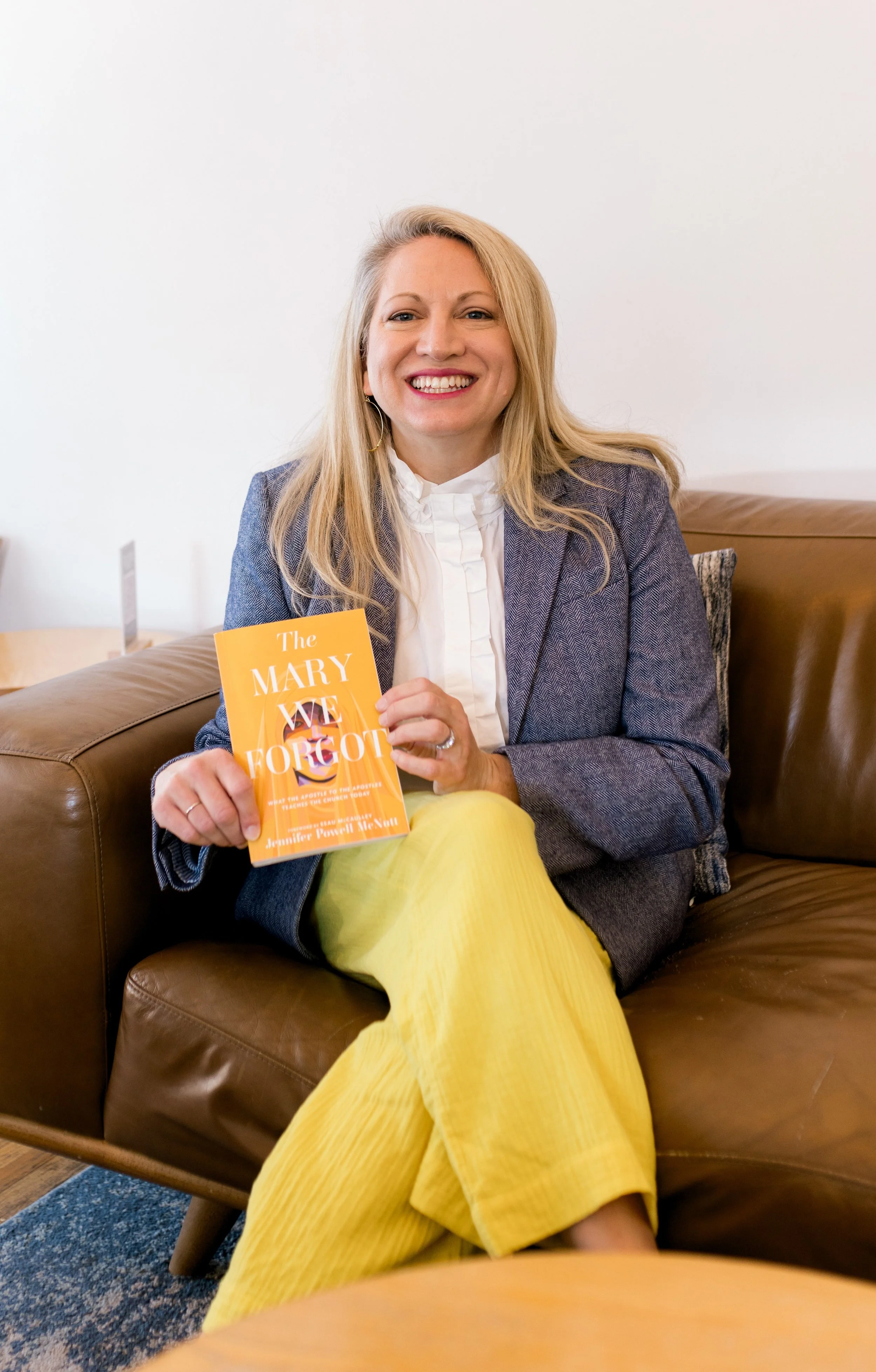 A woman with blonde hair wearing a blazer and white blouse sitting on a brown leather couch, holding a yellow book titled 'The Mary We Forgot.' She is smiling and wearing yellow pants, with a pillow behind her and a white wall in the background.