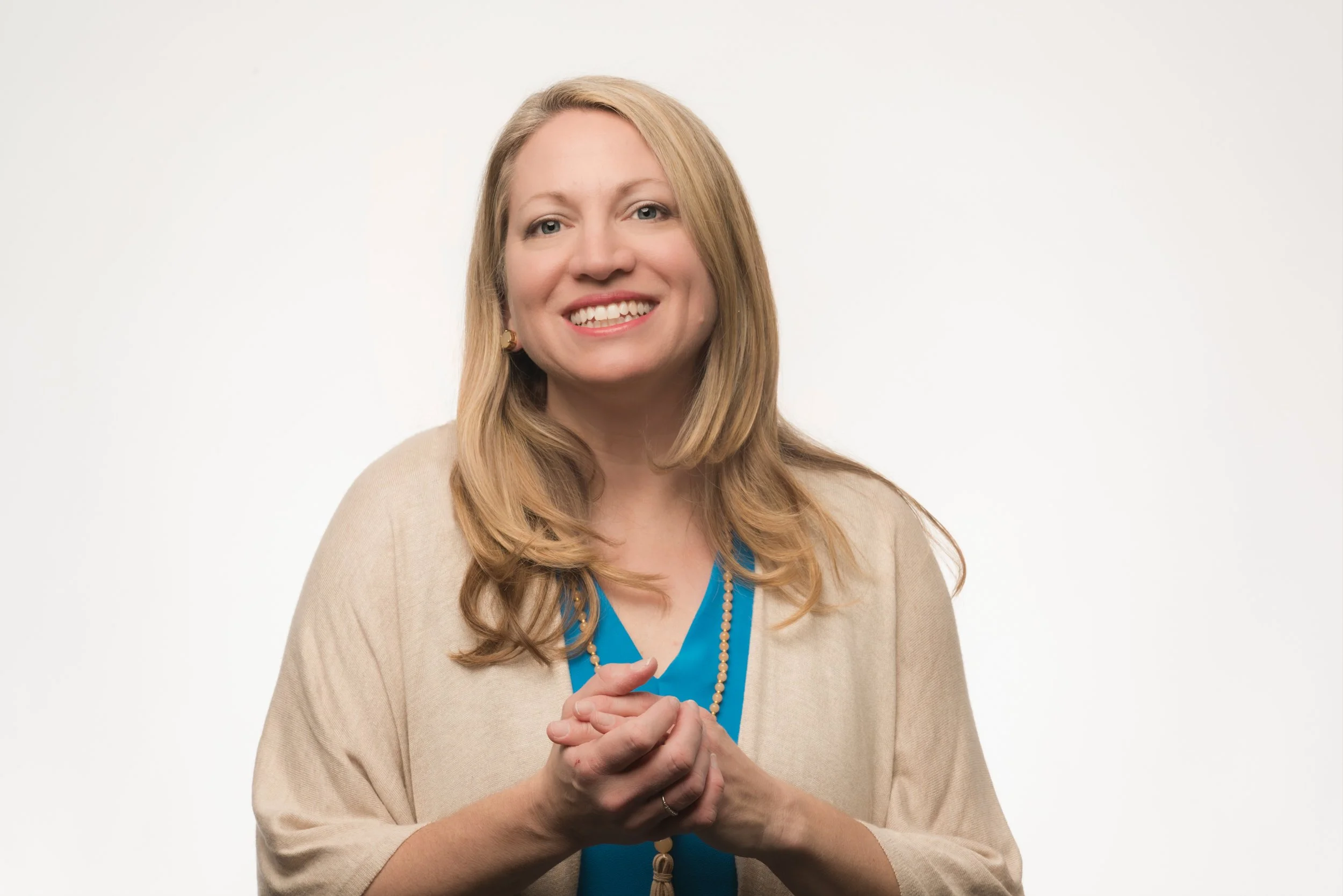 A woman with long, wavy blonde hair smiling at the camera, wearing a beige cardigan over a blue top, with her hands clasped together.