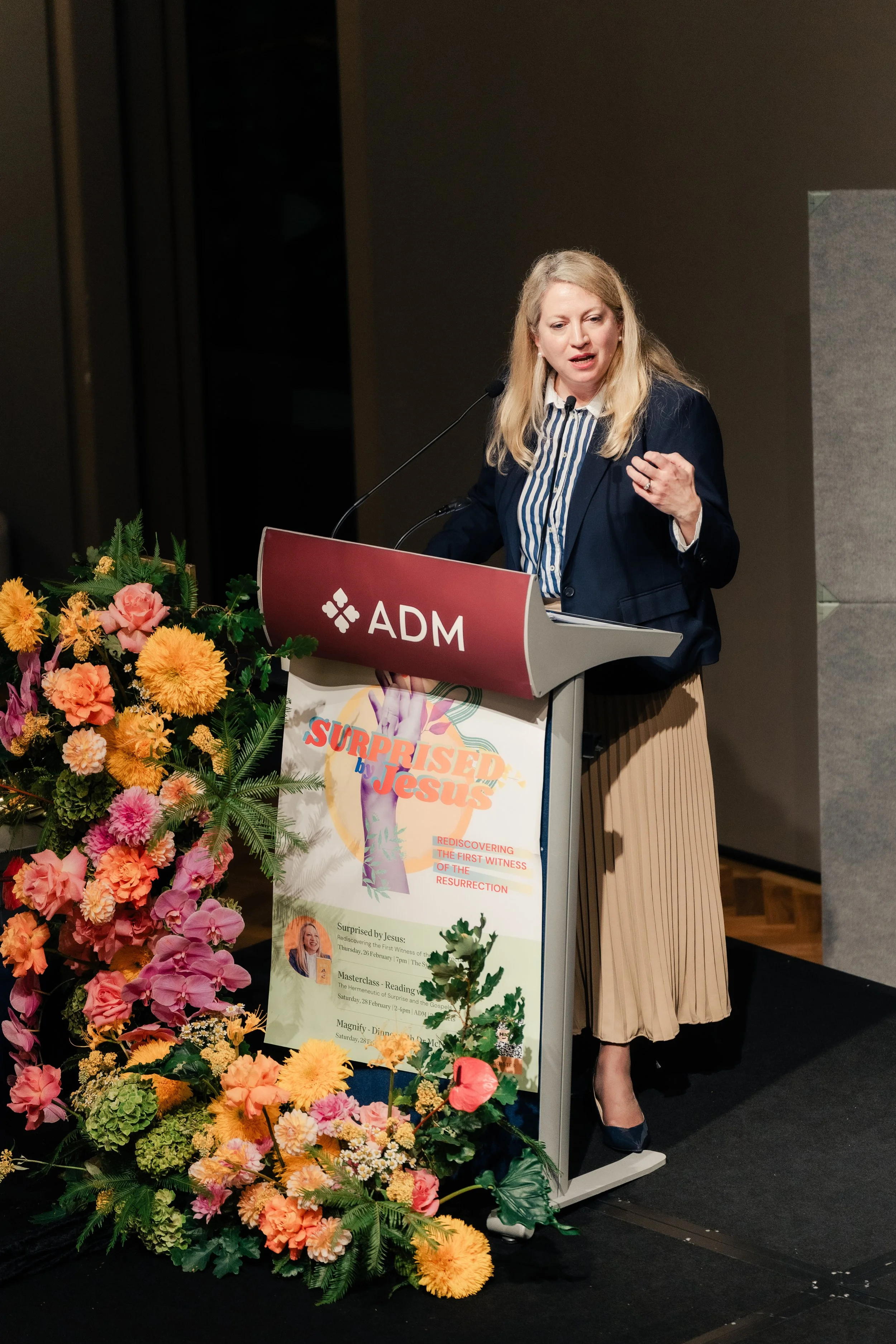 A woman giving a speech at a podium with a sign reading "ADM" and a colorful poster for an event titled "Surprised by Jesus." The podium is decorated with a large arrangement of pink, orange, yellow, and green flowers. The woman is wearing a dark bla