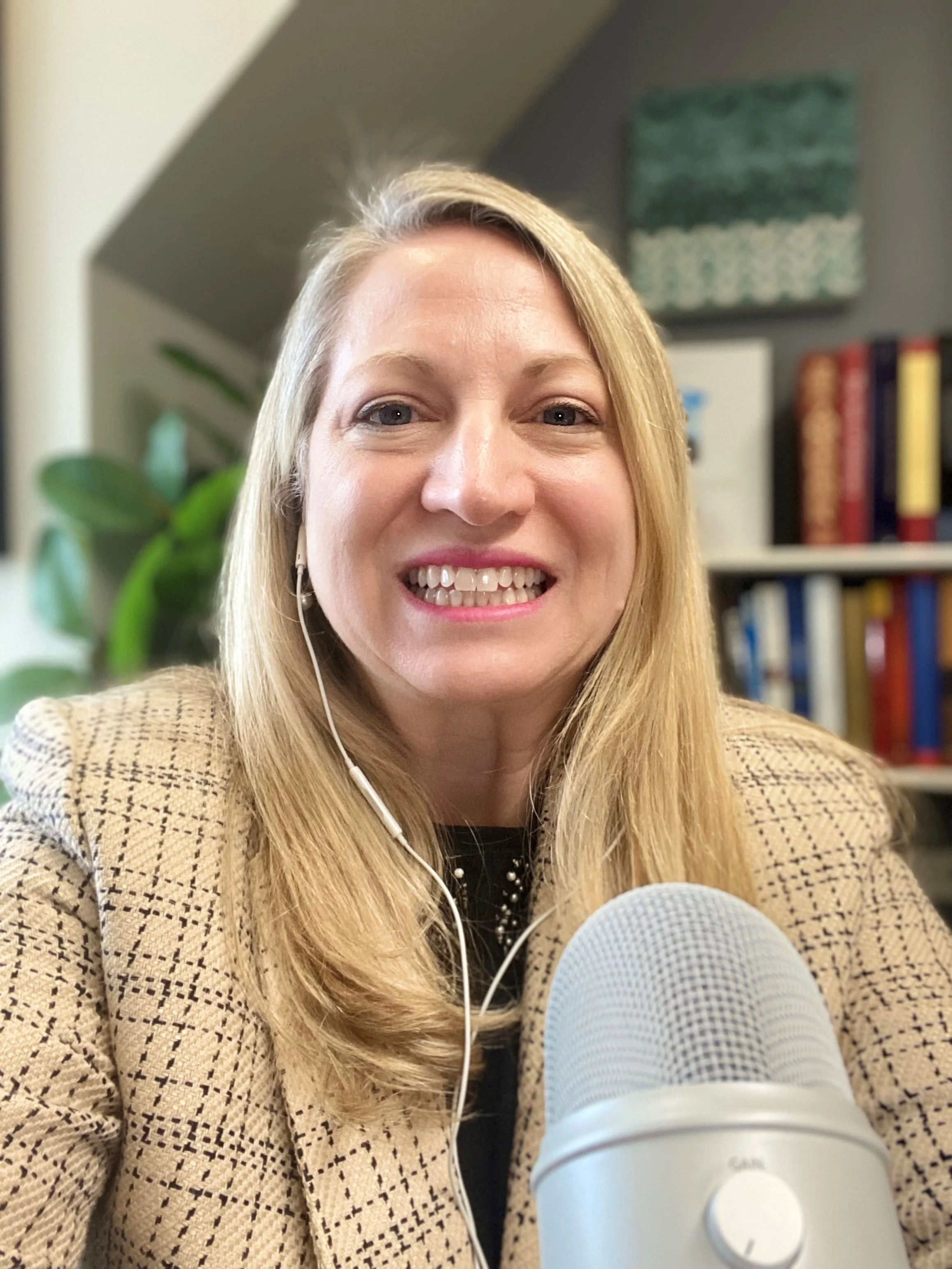 A woman with blonde hair smiling at the camera, wearing a tan checked blazer, with a bookshelf and a green patterned art piece in the background, and a blue microphone in the foreground.