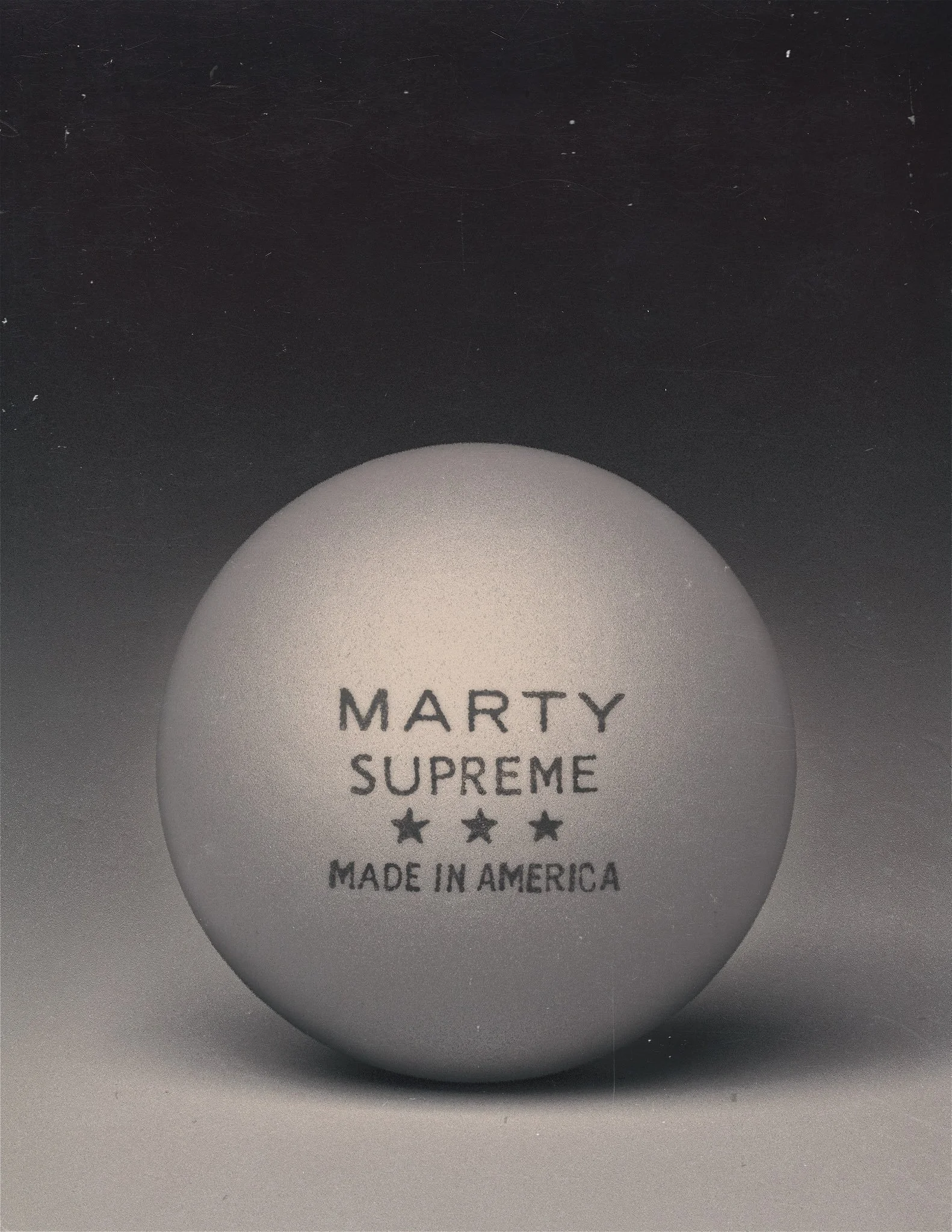 A ping pong ball with the text 'MARTY SUPREME MADE IN AMERICA' and three stars printed on it, set against a dark background.