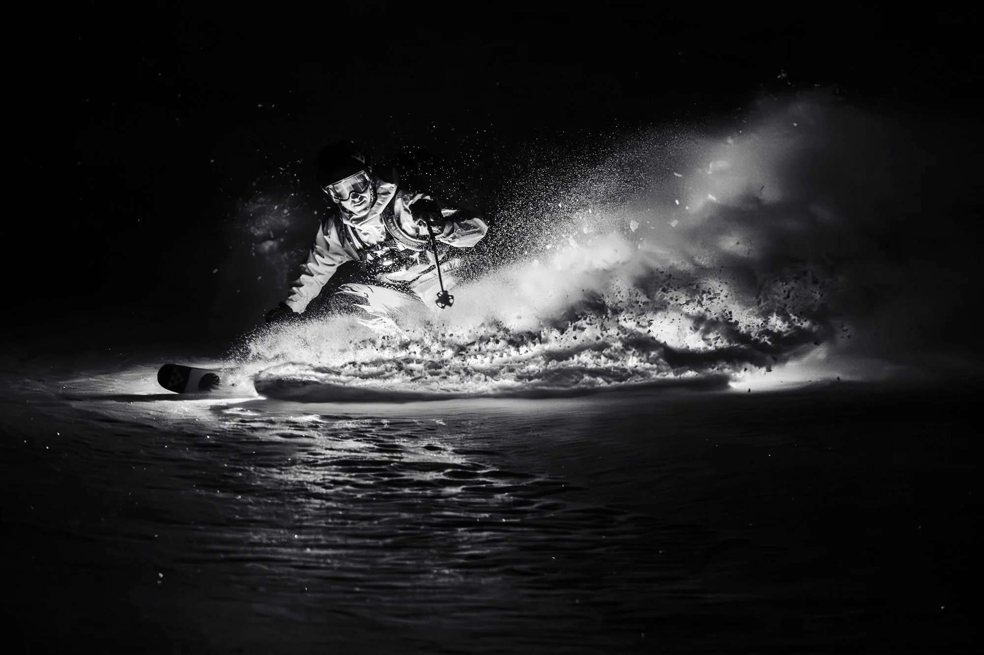 Monochrome photo of a skier at night with a chest mounted light illuminating the snow spray from their skis.