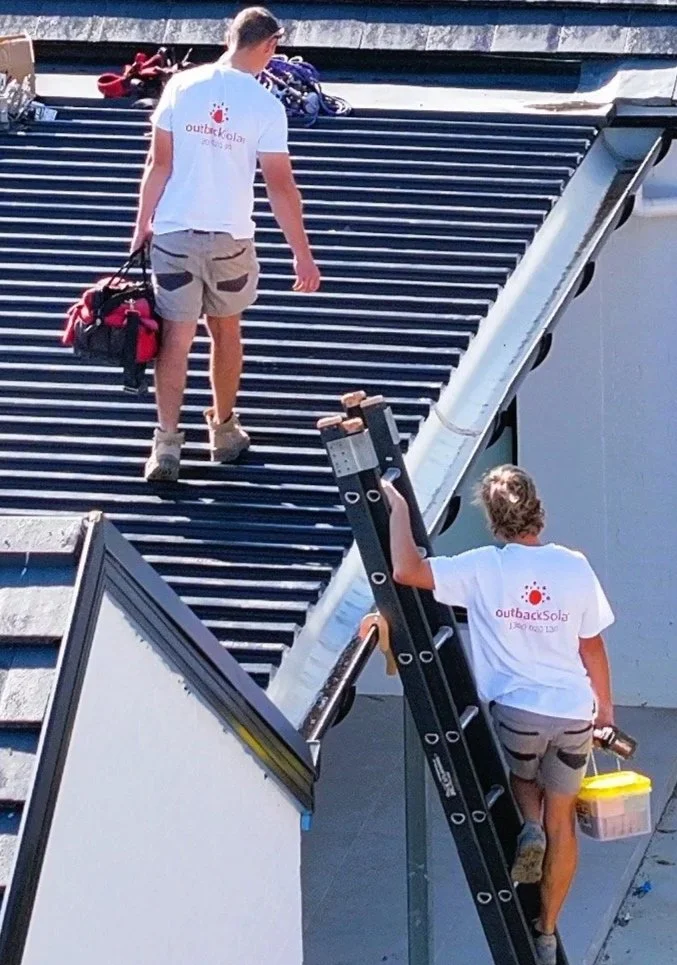 Two men wearing white shirts with 'Outback Solar' logos are walking up a staircase. One man is carrying a toolbox, while the other is holding a red bag and a drink container. They are ascending metal stairs outside, with solar panels on the roof abov
