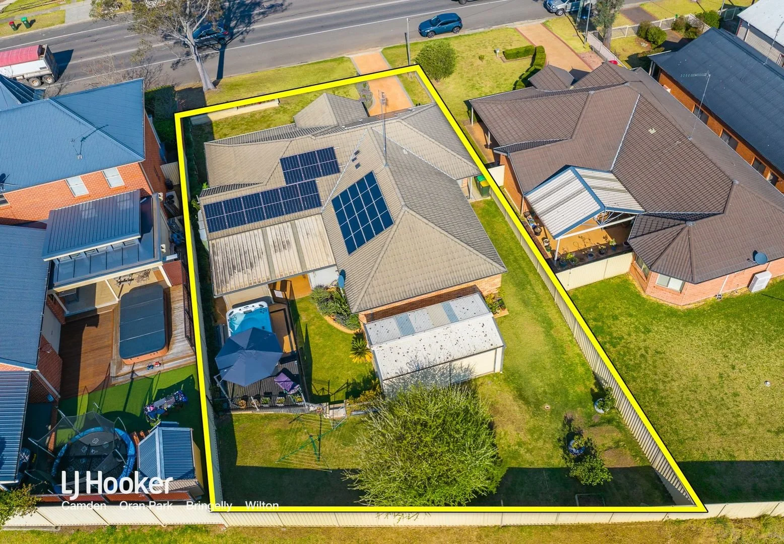 Aerial view of a backyard with a large house equipped with solar panels, a kiddie pool, a shaded patio area, a shed, and a green lawn, enclosed by a yellow boundary fence.