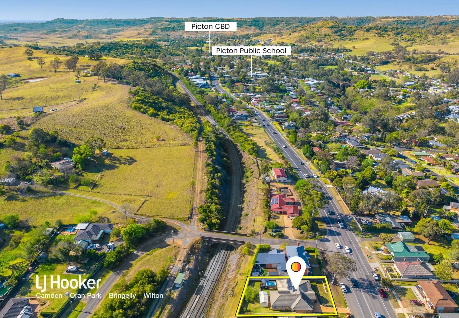 Aerial view of a suburban neighborhood with residential houses, a railway, and nearby green hills. Labels indicate Picton CBD, Picton Public School, and the image is from LJ Hooker Camden, Oran Park, Bringelly, Wilton.