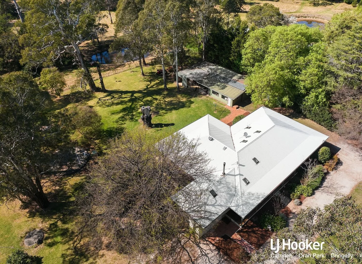 Aerial view of a house with a white roof, surrounded by large trees, green lawn, and a small brick pathway leading to a side door. There is a large tree trunk stump on the lawn and a small shed or outbuilding with a green roof nearby. The property is in a semi-rural area with open land and a pond visible in the background.