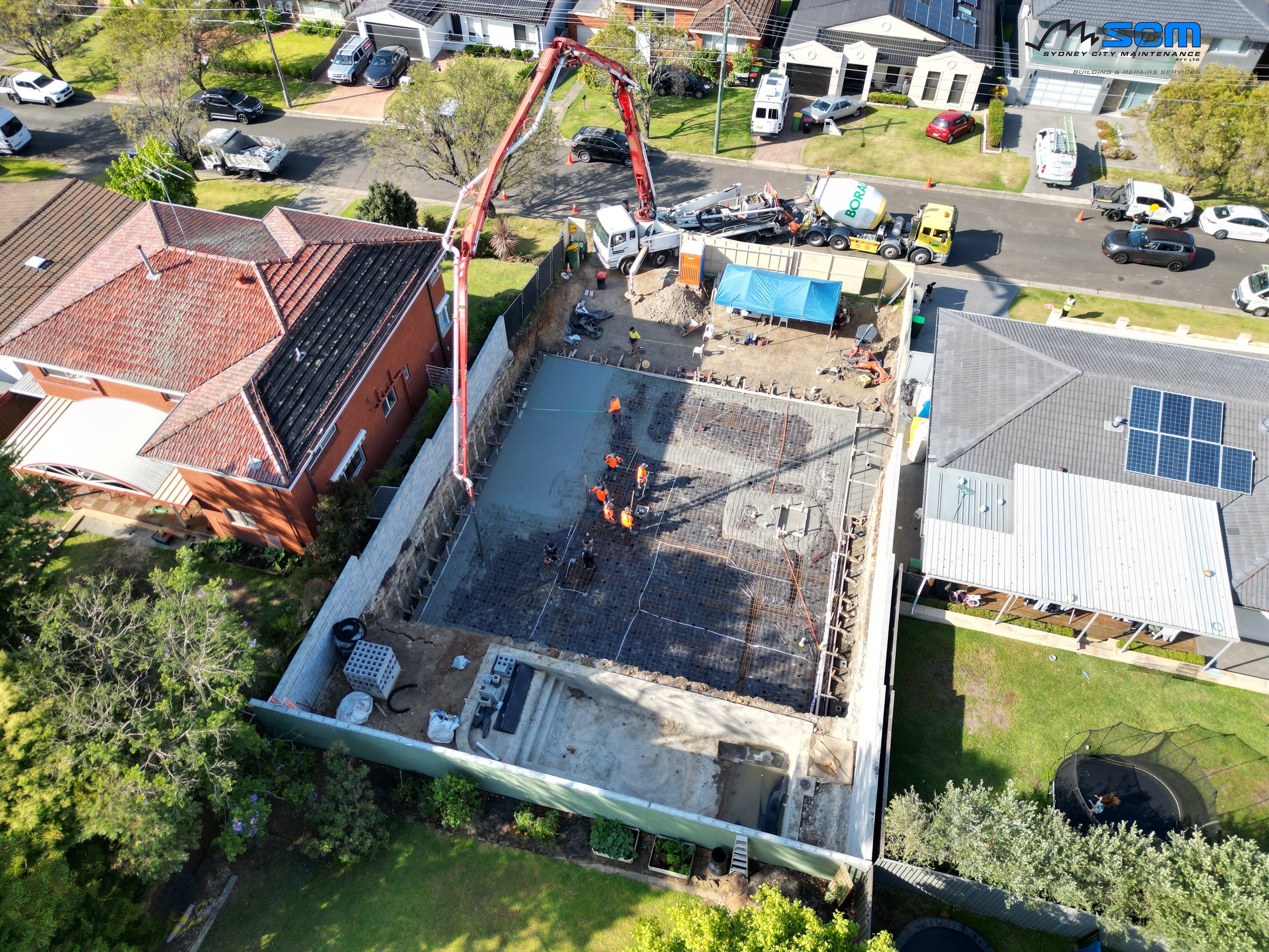 A construction site in a residential neighborhood where workers pour concrete for a building's foundation. A concrete pump truck is pouring concrete into the foundation, and construction workers in orange vests are working on the site. Adjacent house