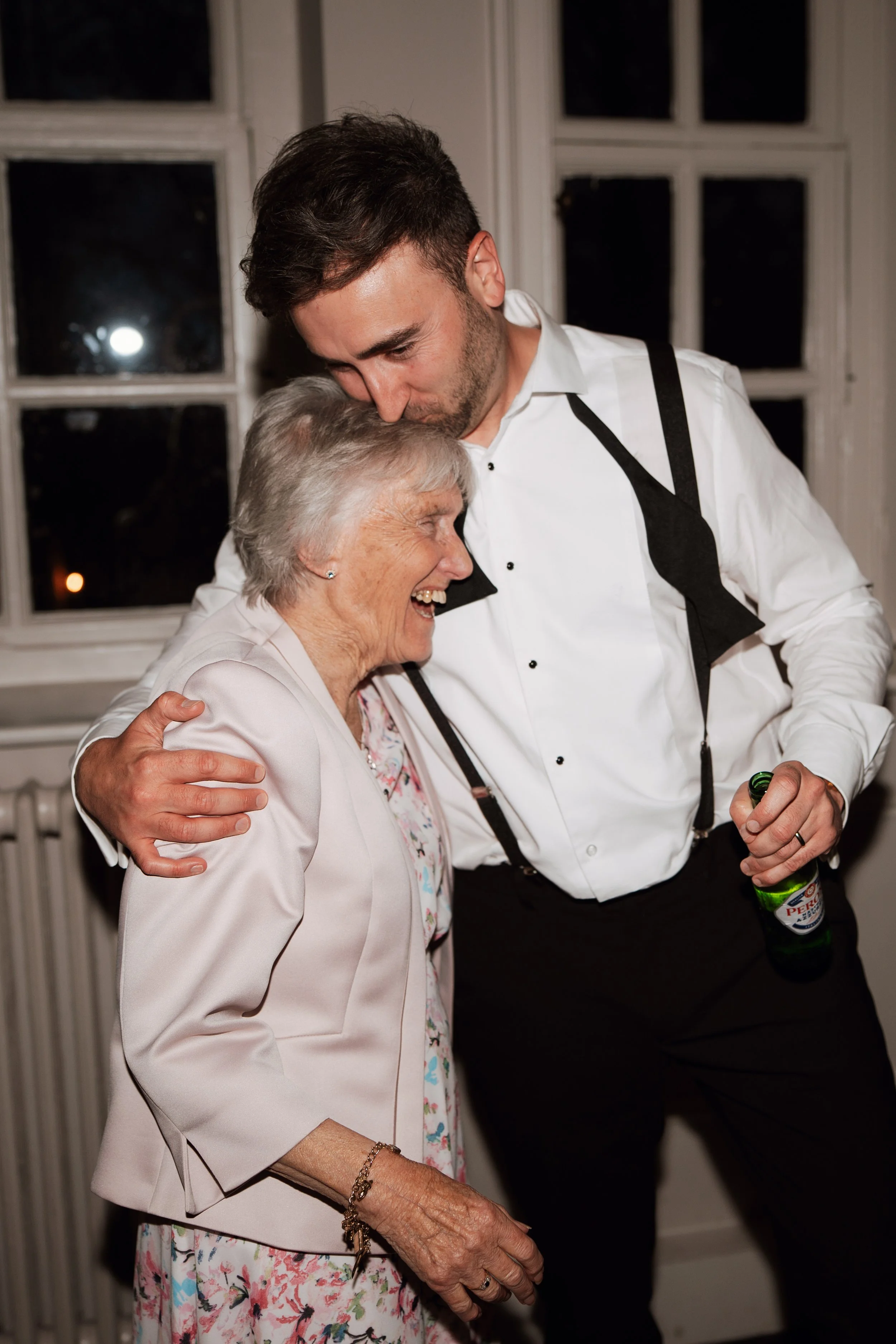A young man and an elderly woman sharing a warm hug, both smiling, inside a room with windows and white walls at Howsham hall 